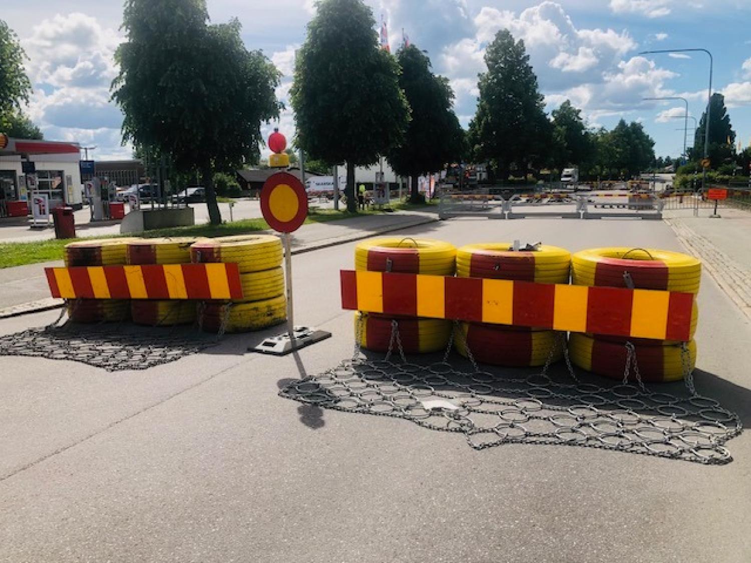 Gatan är avspärrad med röda och gula avspärrningsbarriärer, en skylt med ”Ingen infart” och kedjor, vilket tyder på att vägen är avstängd. I bakgrunden syns träd och byggnader under en delvis molnig himmel.