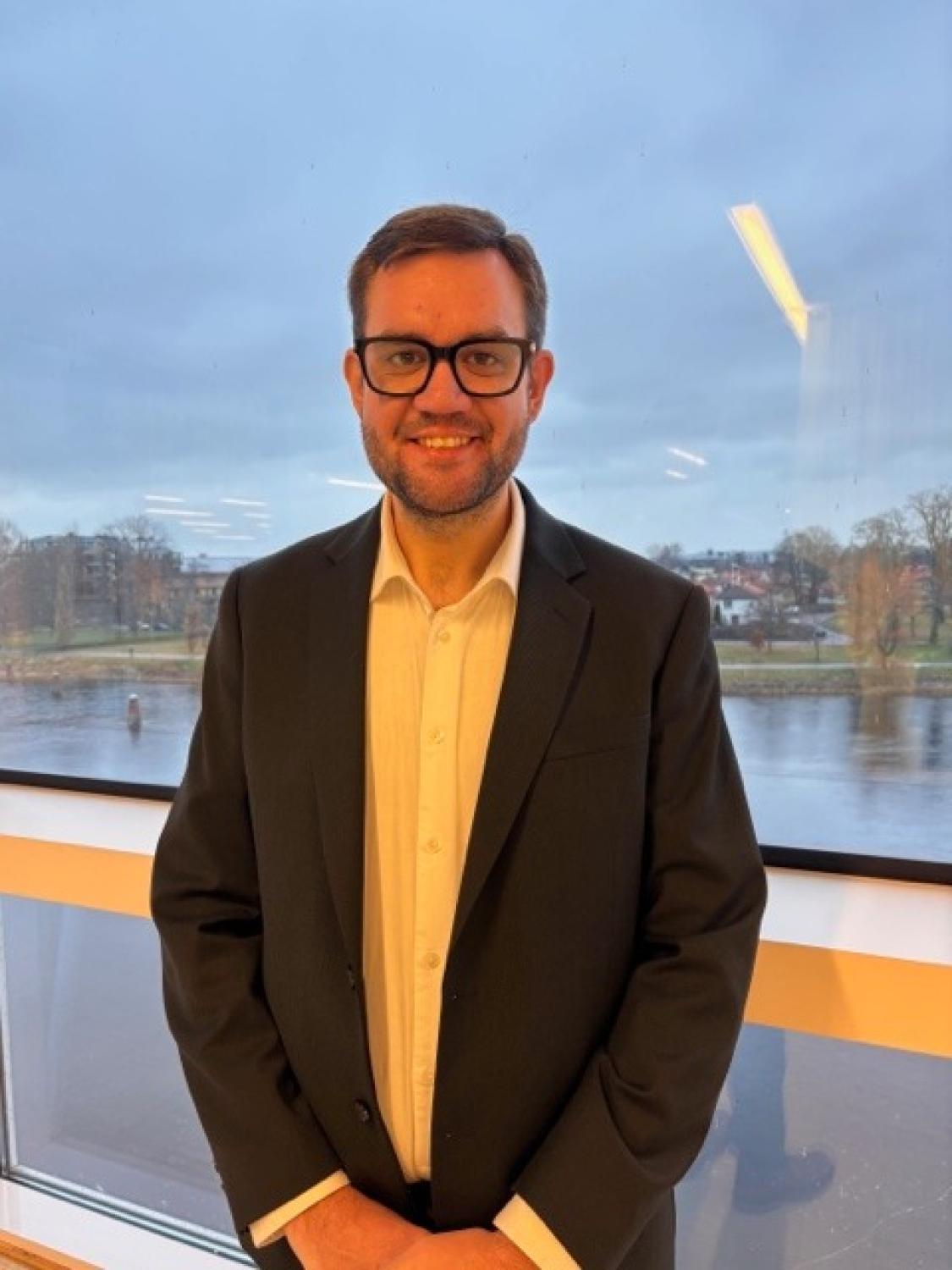 En man i kostym och glasögon står inomhus vid ett stort fönster med en flod och träd som syns utanför en molnig dag.
