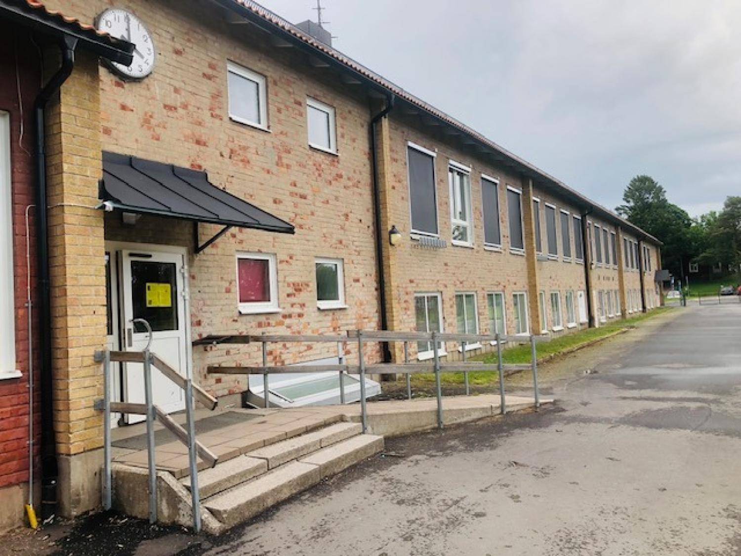En tvåvåningsbyggnad i tegel med stora fönster, en ramp och trappsteg vid ingången samt en klocka ovanför dörren. Marken ser våt ut, vilket tyder på att det nyligen har regnat.