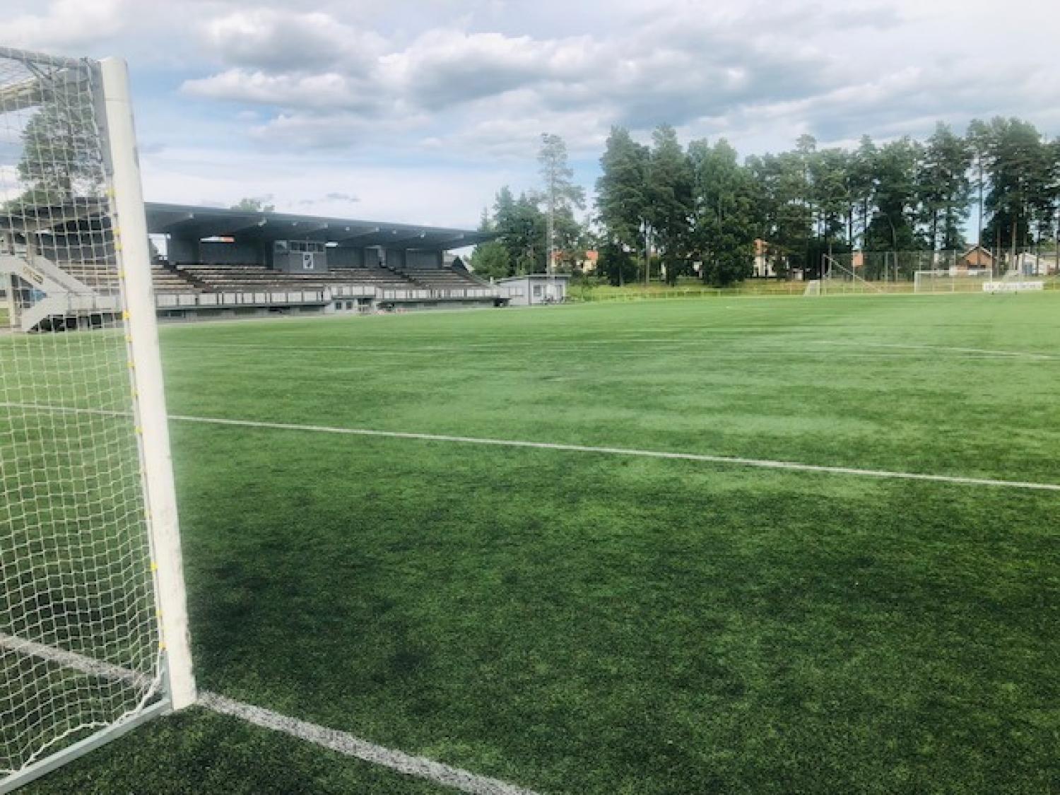 En fotbollsplan med konstgräs, sedd bakifrån en målstolpe, med tomma läktare och träd i bakgrunden under en molnig himmel.