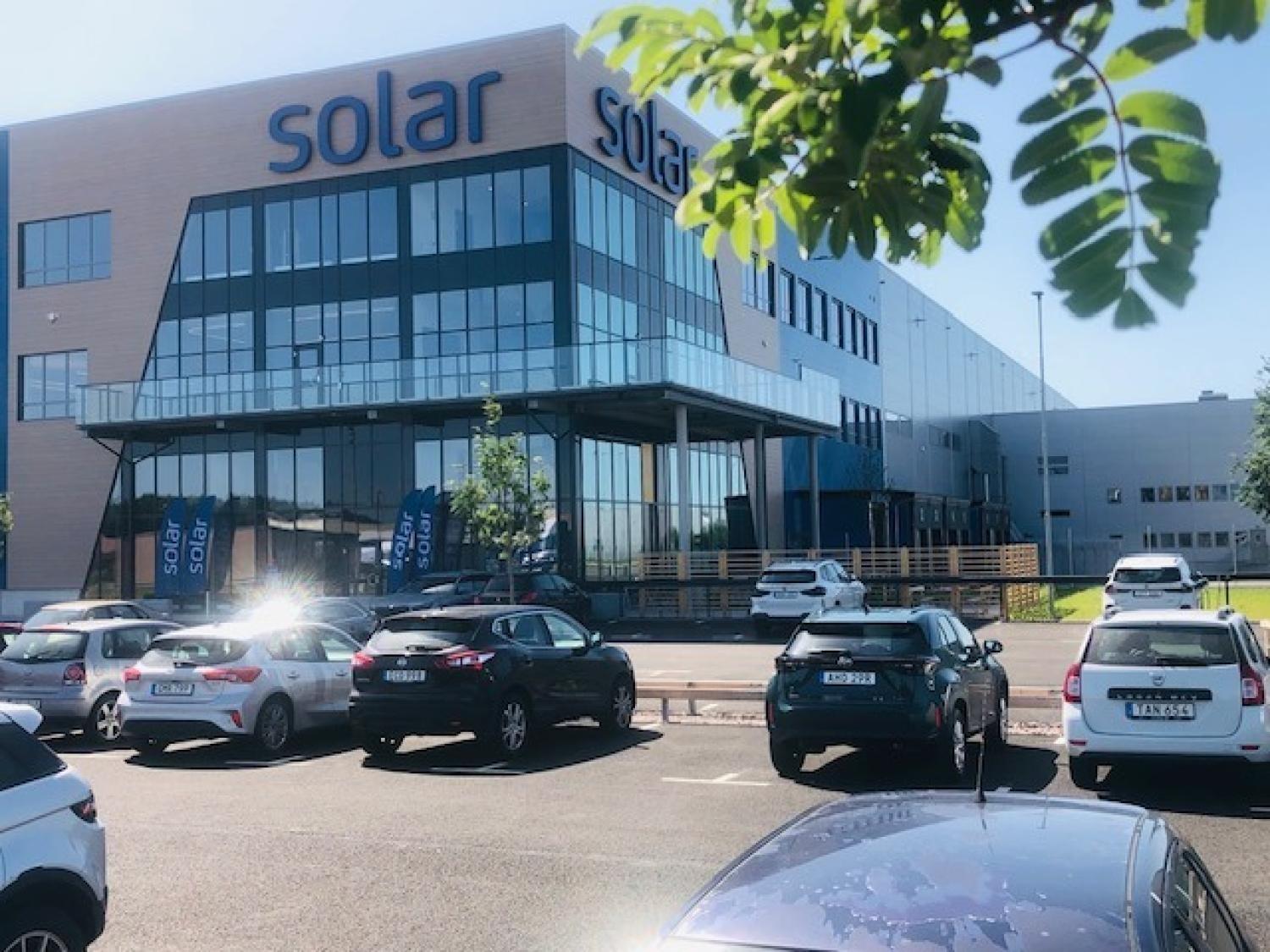 En modern kontorsbyggnad med skyltar med solmotiv, stora glasfönster och en parkeringsplats full av bilar under en klar himmel.