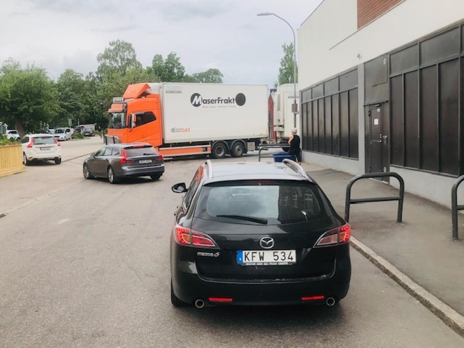 En svart Mazda med svenska registreringsskyltar står parkerad på gatan nära trottoaren, medan en lastbil och en grå sedan står längre fram, och en person står i närheten.