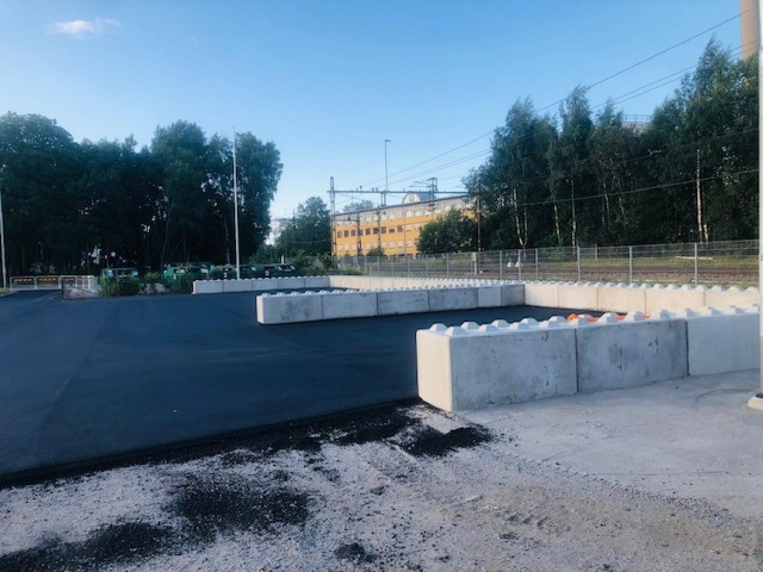 En nyasfalterad yta med betongbarriärer, omgiven av träd och staket, med en gul byggnad i bakgrunden under en klar himmel.