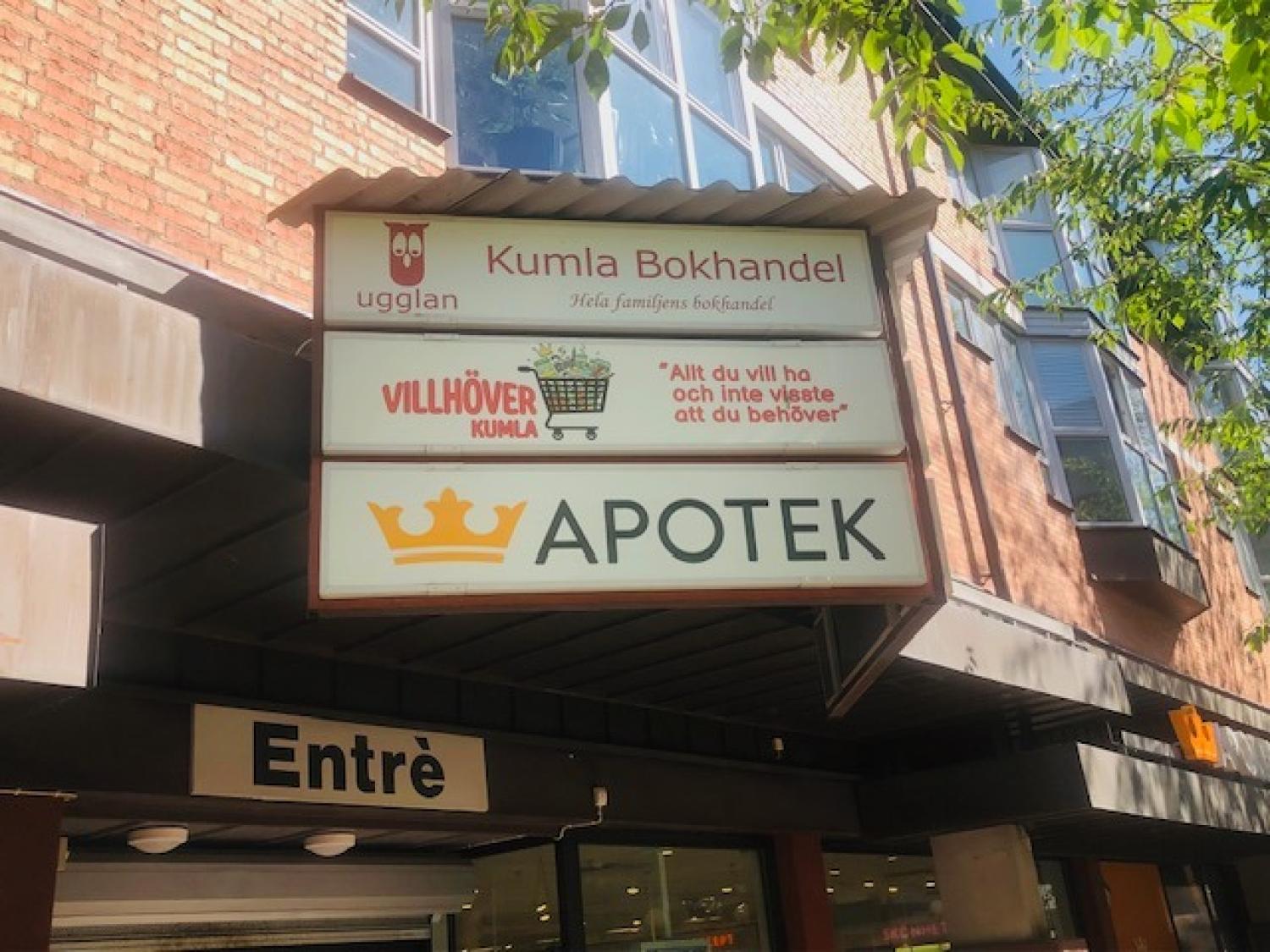 En butiksskylt för Kumla Bokhandel och Apotek på en tegelbyggnad, där träden delvis skuggar skyltarna.