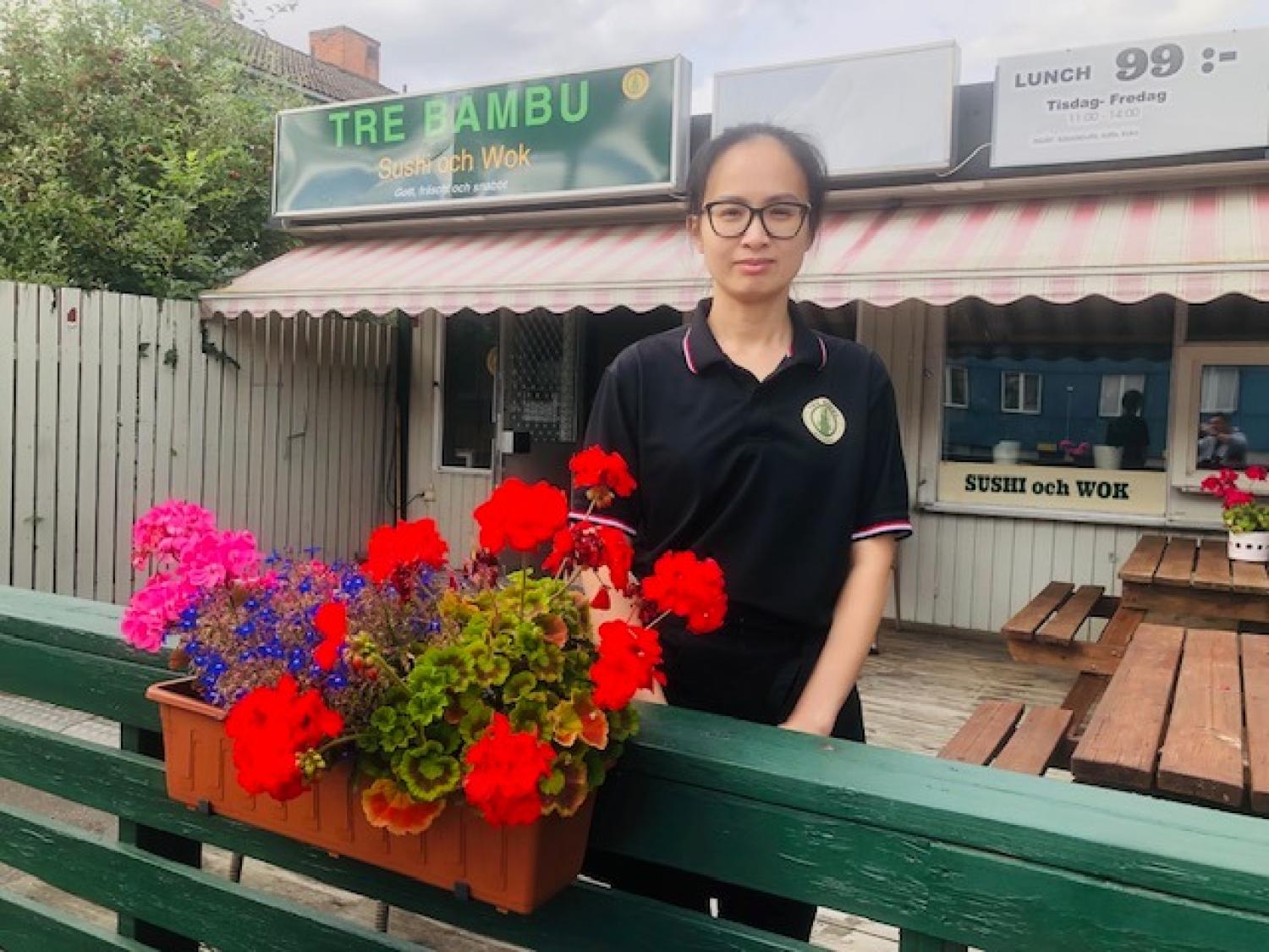 En kvinna i svart uniform står vid ett grönt staket med en blomlåda framför en restaurang som heter Tre Bambu, och bakom henne syns uteserveringen.