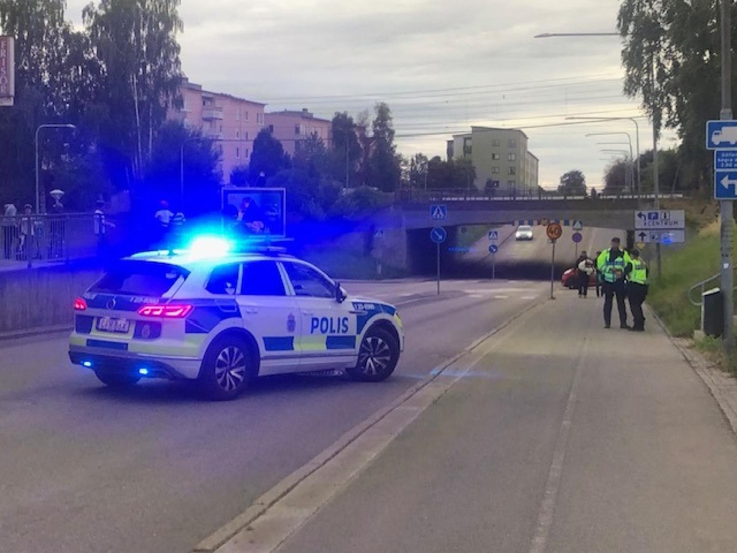 En polisbil med blå blinkljus spärrar av en väg nära en tunnel, medan två poliser står på trottoaren intill.