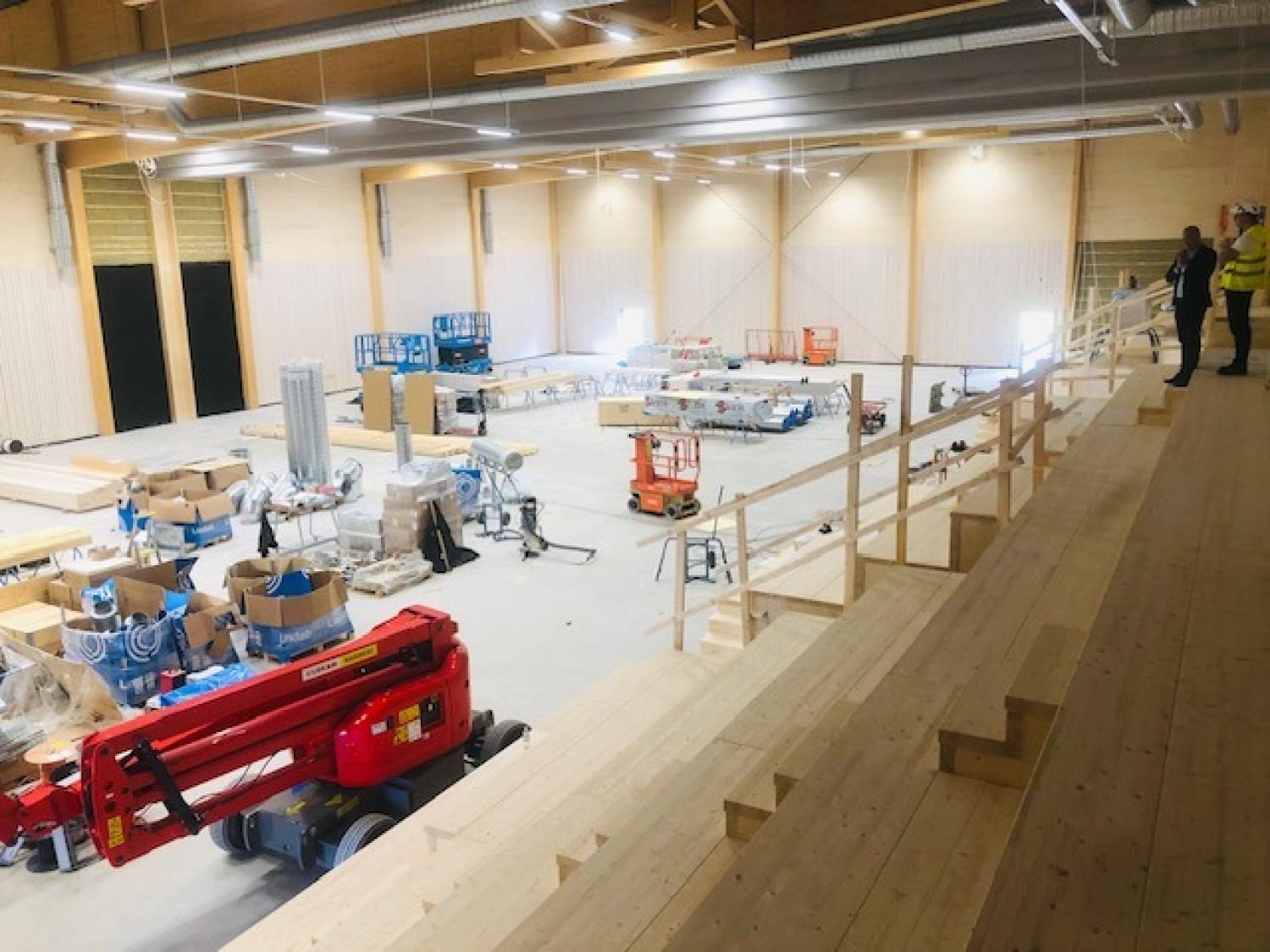 En stor byggarbetsplats inomhus med läktare av trä, utrustning, byggmaterial och två personer i skyddsutrustning som observerar från en högre nivå.