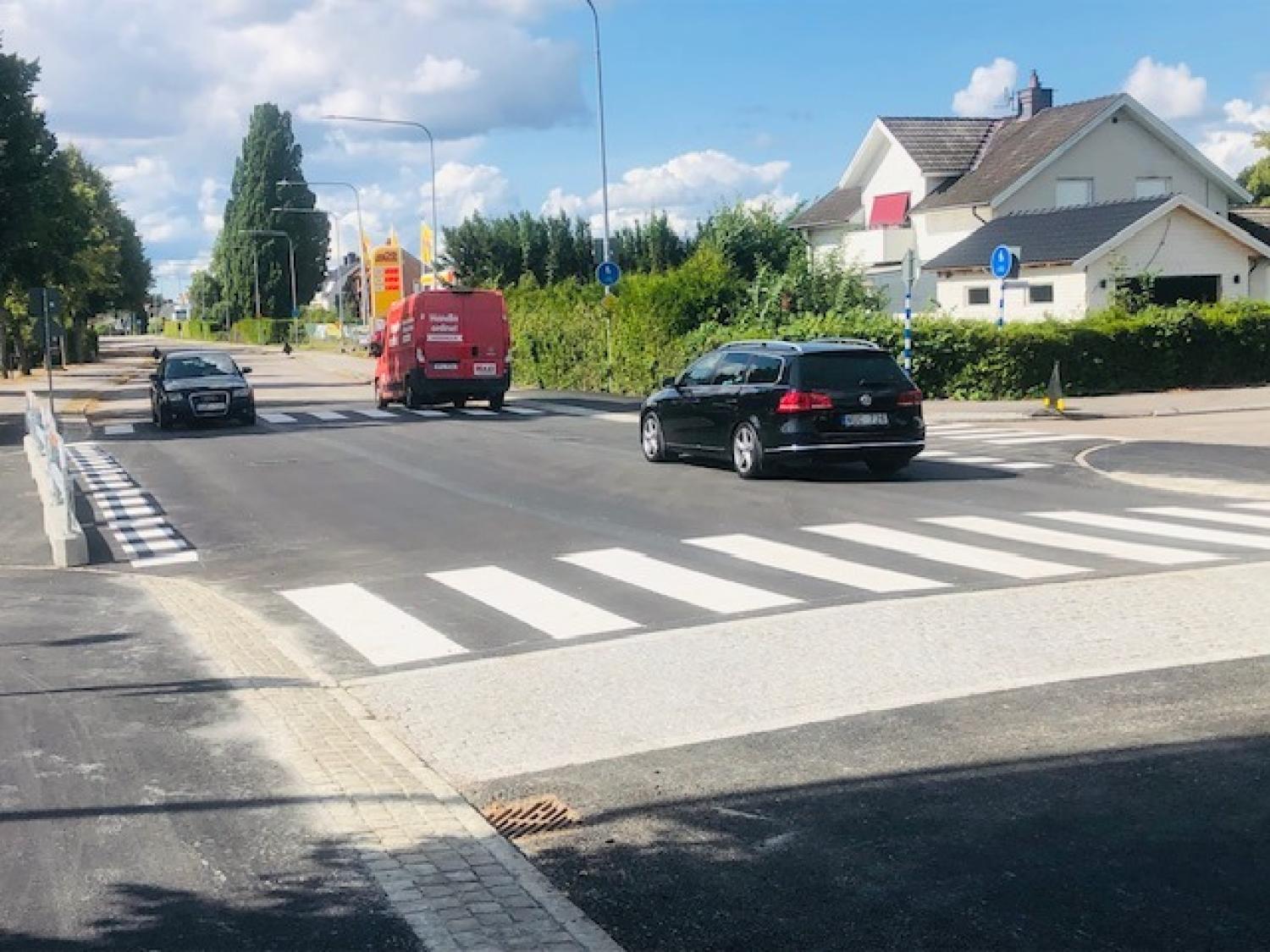 Tre fordon, däribland en röd skåpbil och två personbilar, kör på en asfalterad väg med övergångsställen i ett förortsområde en solig dag.
