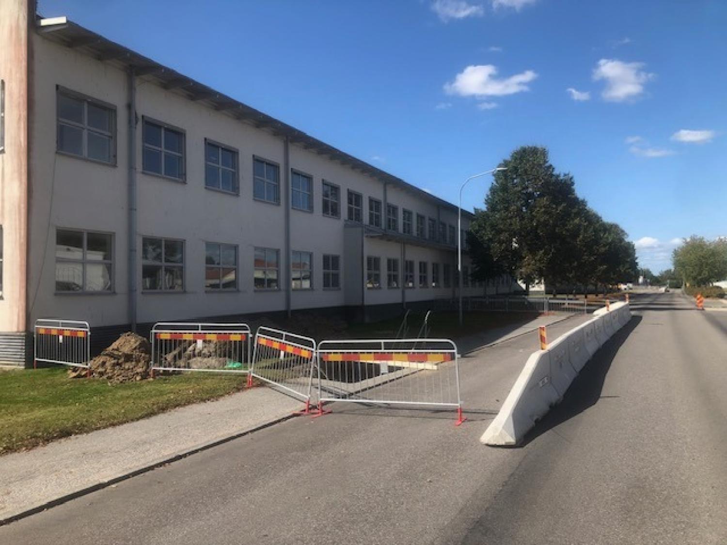 En byggstängsel spärrar av en del av trottoaren nära en byggnad, med trafikavspärrningar längs den intilliggande vägen under en blå himmel.