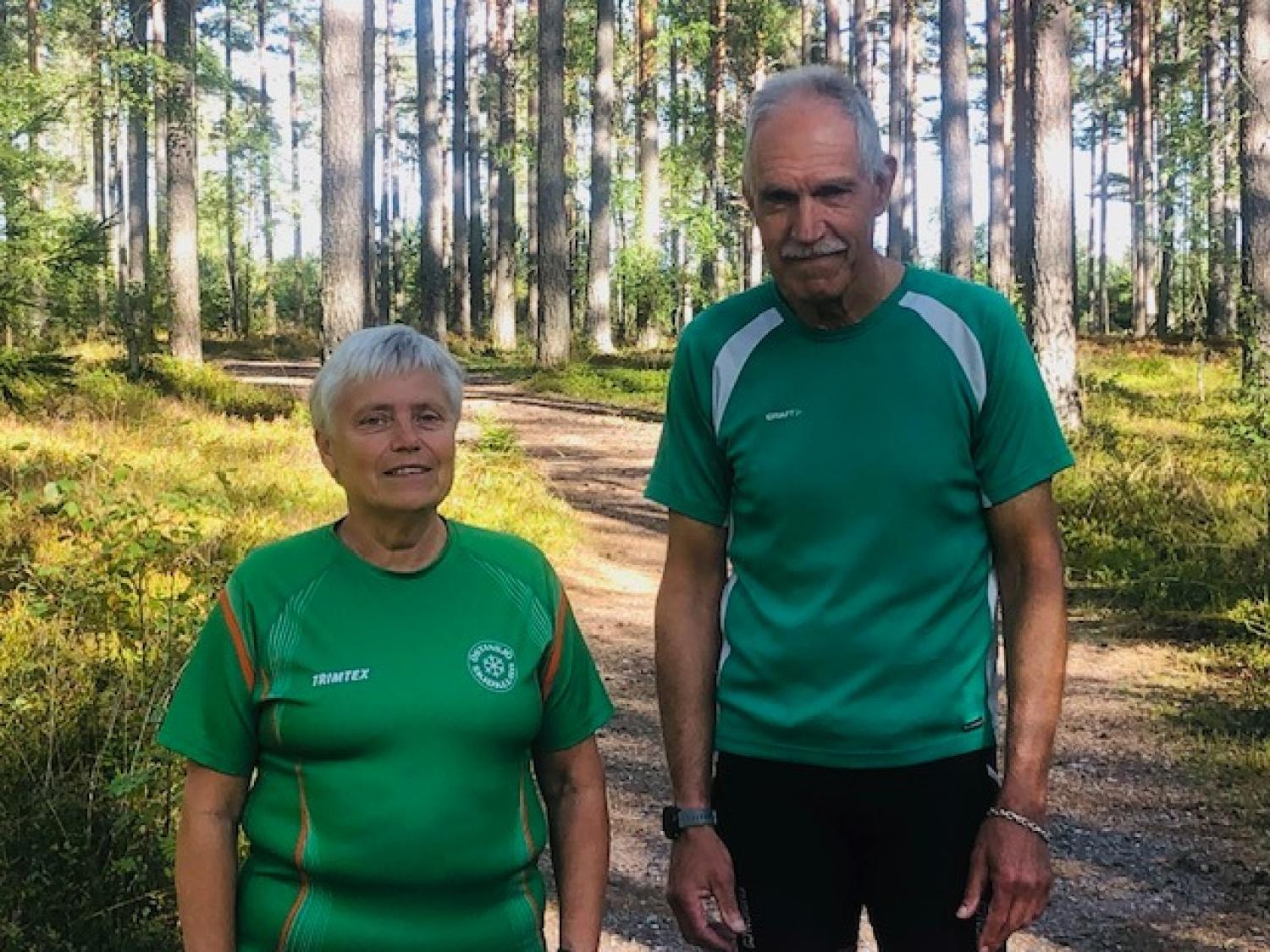 Två äldre personer står på en solbelyst skogsstig, båda klädda i gröna träningströjor. De är omgivna av träd och grönska.