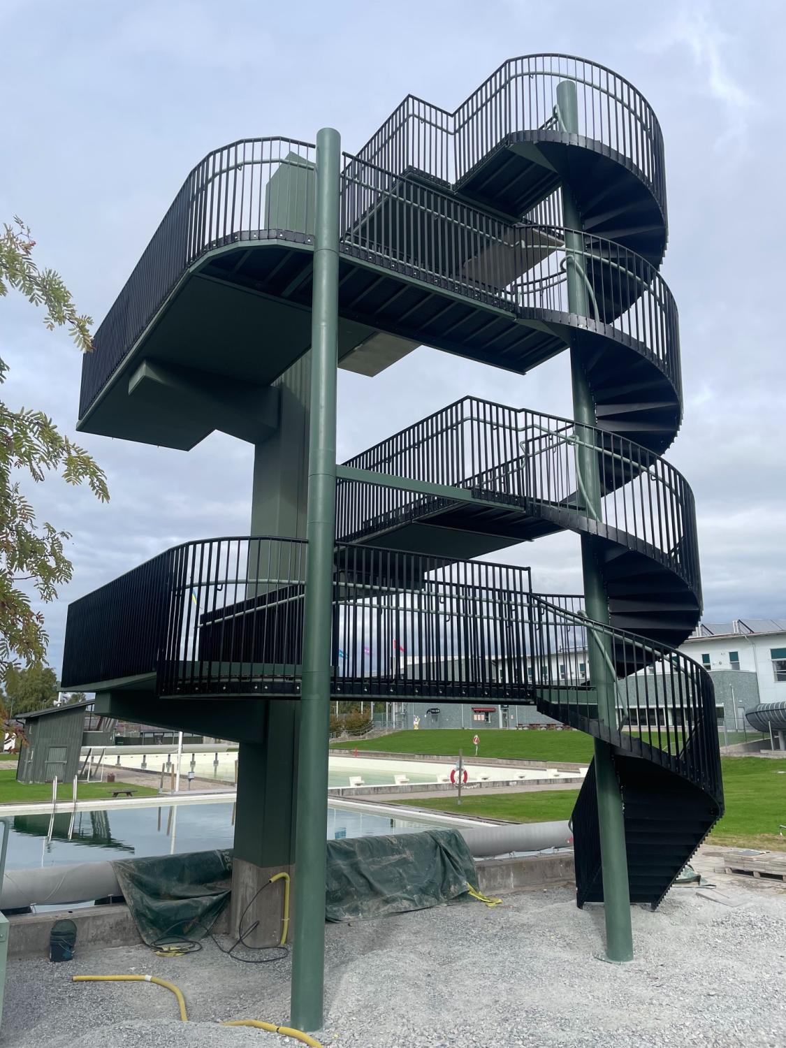 En hög spiraltrappa med svarta räcken leder upp till en upphöjd plattform som ligger utomhus nära ett poolområde och är omgiven av byggmaterial.