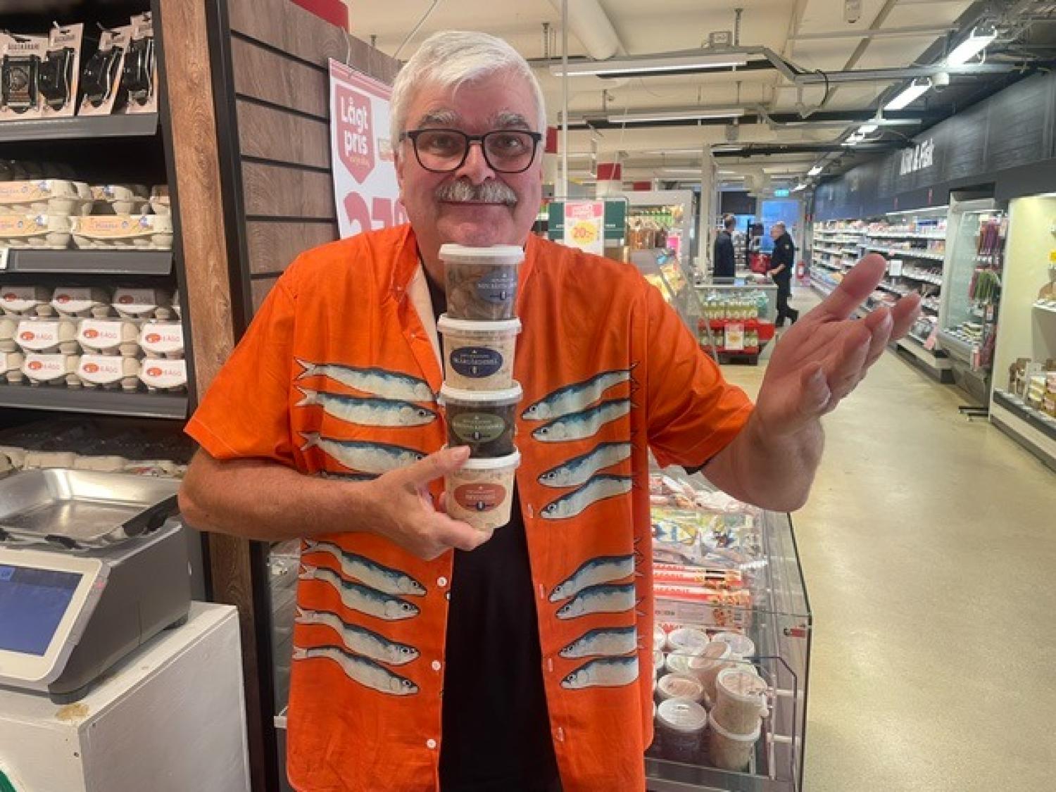 En man i en orange skjorta med fiskmönster står i en gång i en livsmedelsbutik med en stapel bestående av fem små matförpackningar i händerna och ler mot kameran.
