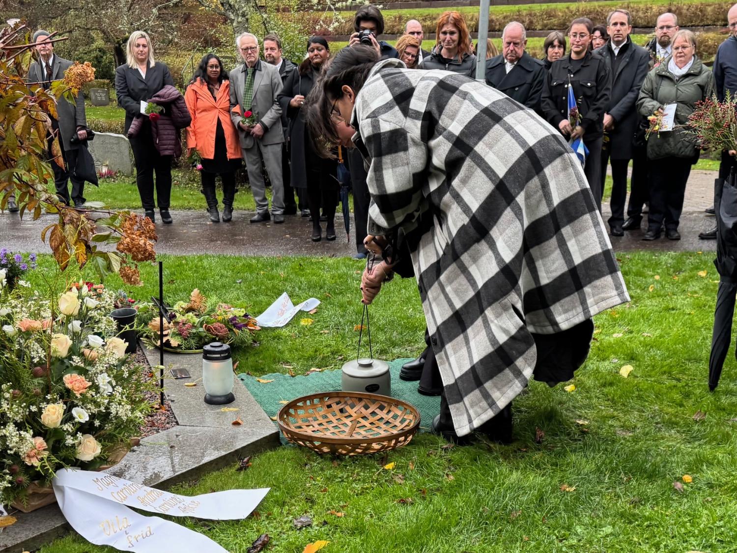 En grupp människor i formell klädsel samlas vid en gravplats, medan en person i rutig rock tänder ett ljus nära blomsterarrangemang.