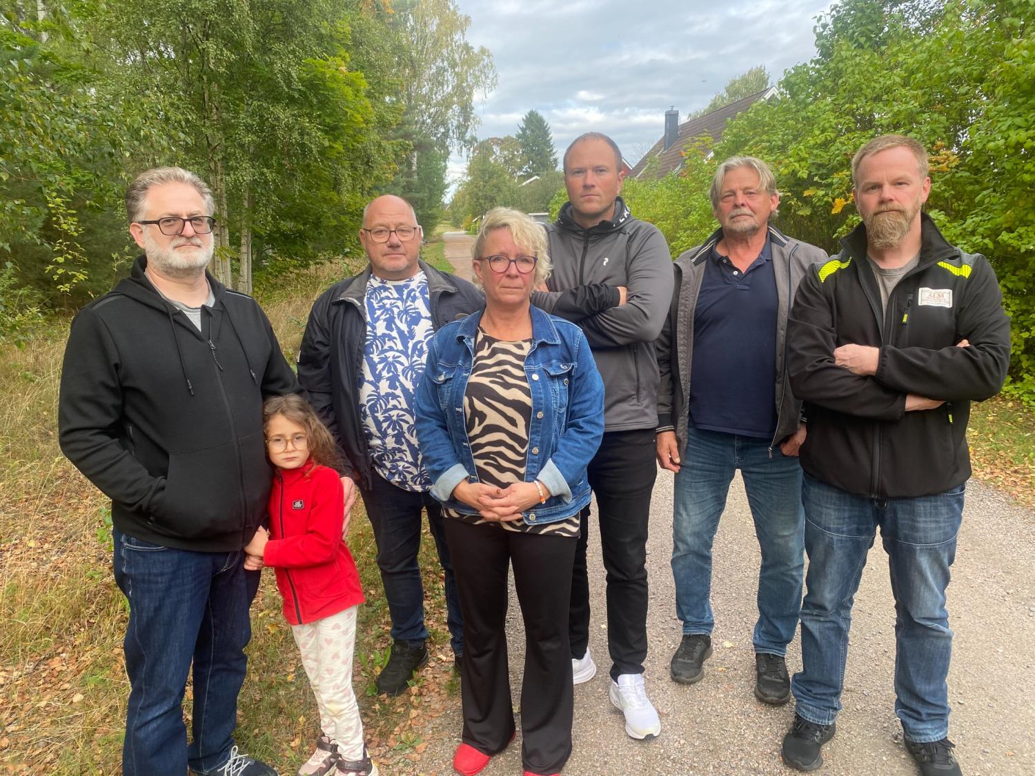 Sju vuxna och ett barn står tillsammans på en trädkantad stig och tittar mot kameran med uttryckslösa ansikten.