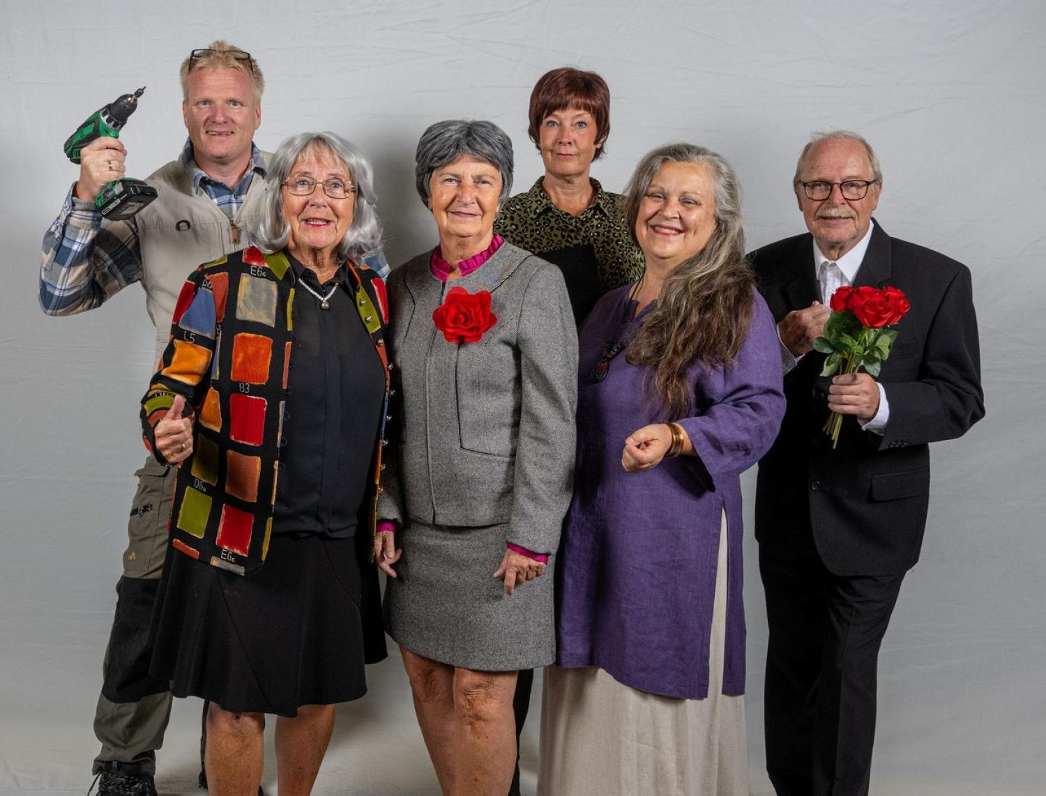 Sex vuxna står och poserar för ett gruppfoto mot en vanlig bakgrund; en håller i en borr, en annan håller i en bukett röda rosor och flera har färgglada kläder och leenden.