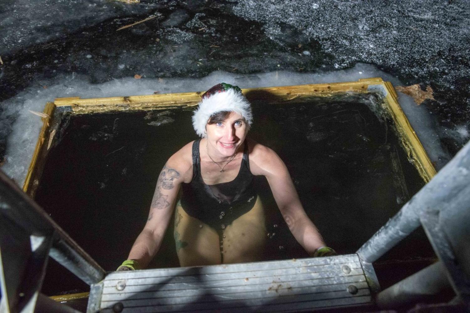 En kvinna i tomteluva och baddräkt står i ett ishål och håller sig fast i en metallstege, med snö och is runt omkring sig.