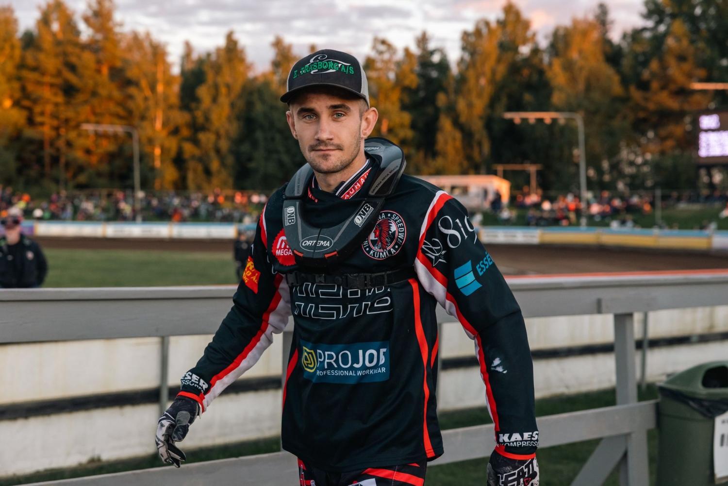 En man i racerdräkt och keps står utomhus nära en racerbana, med en folkmassa och träd i bakgrunden.