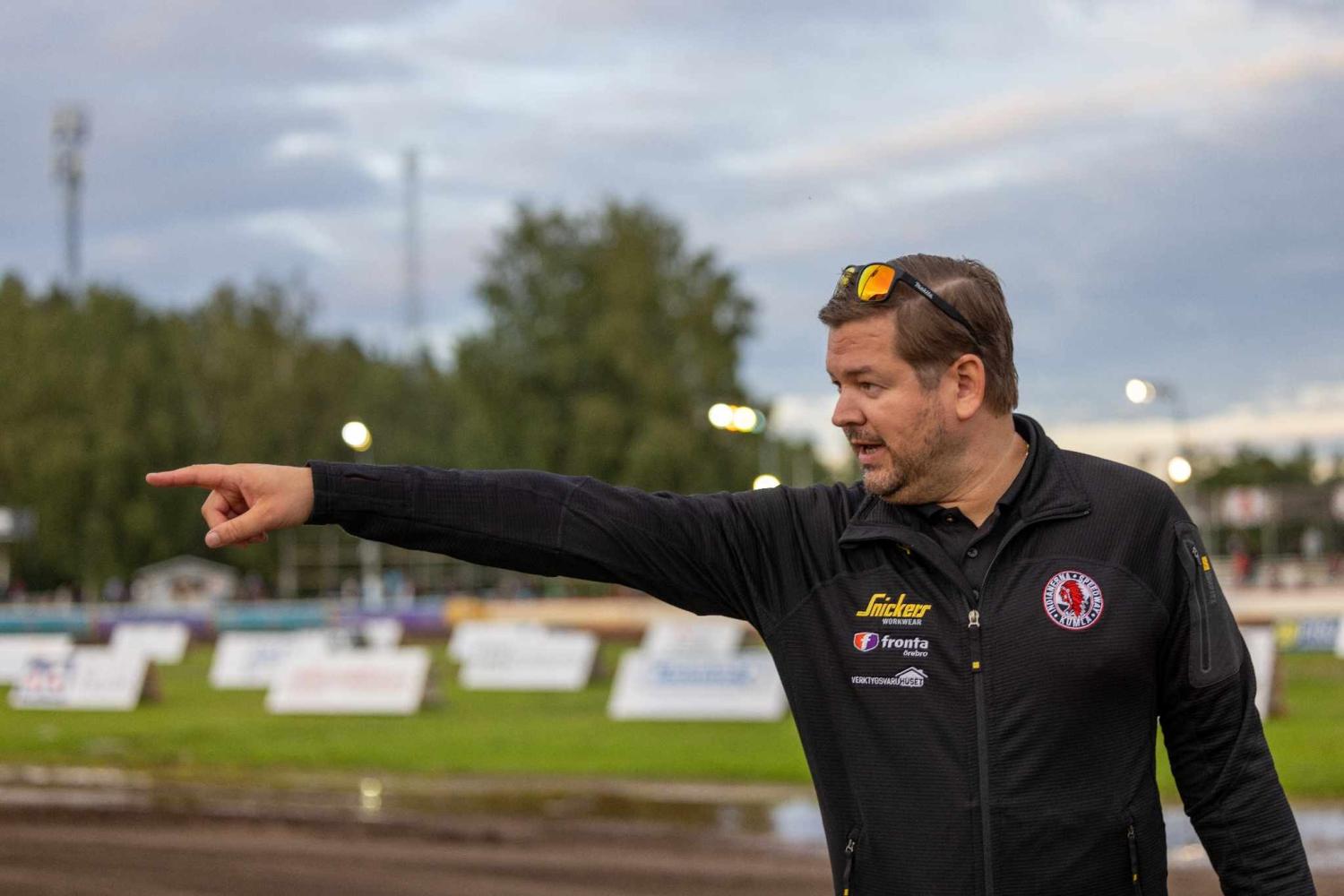 En man klädd i en svart jacka och med solglasögon på huvudet står utomhus och pekar åt vänster, med en suddig bild av en racerbana och träd i bakgrunden.