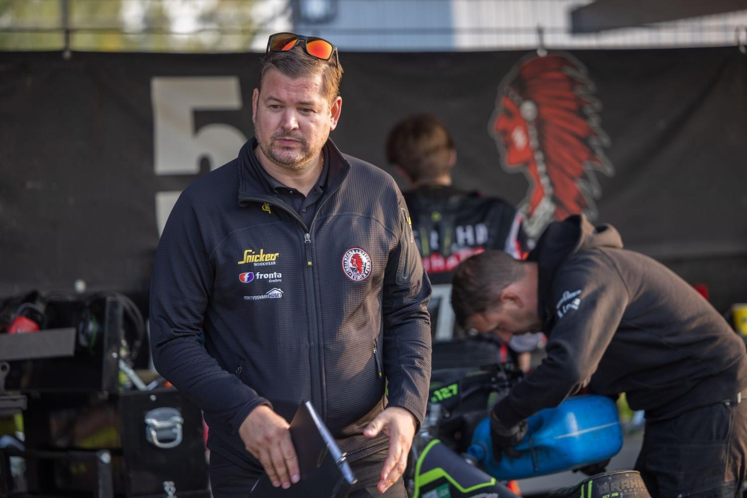 En man i svart jacka håller en anteckningsbok i ett garage, medan en annan person arbetar med utrustning i bakgrunden.