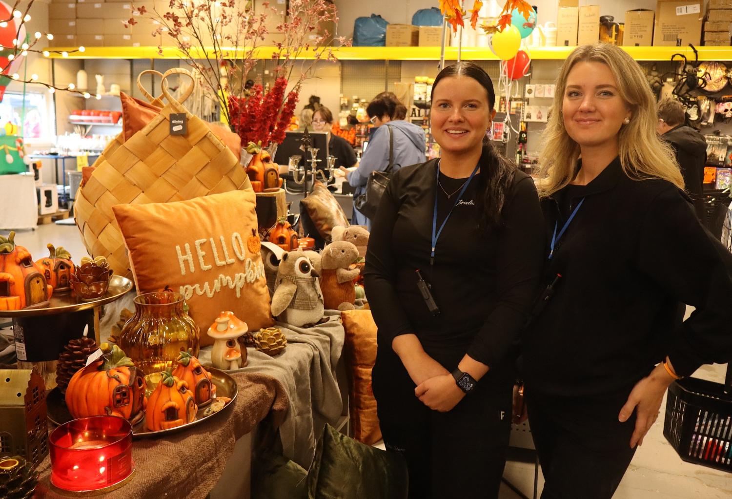 Två kvinnor står bredvid en utställning med höstinspirerade dekorationer, bland annat pumpor, ljus och en kudde med texten ”Hello Pumpkin”, inne i en butik.