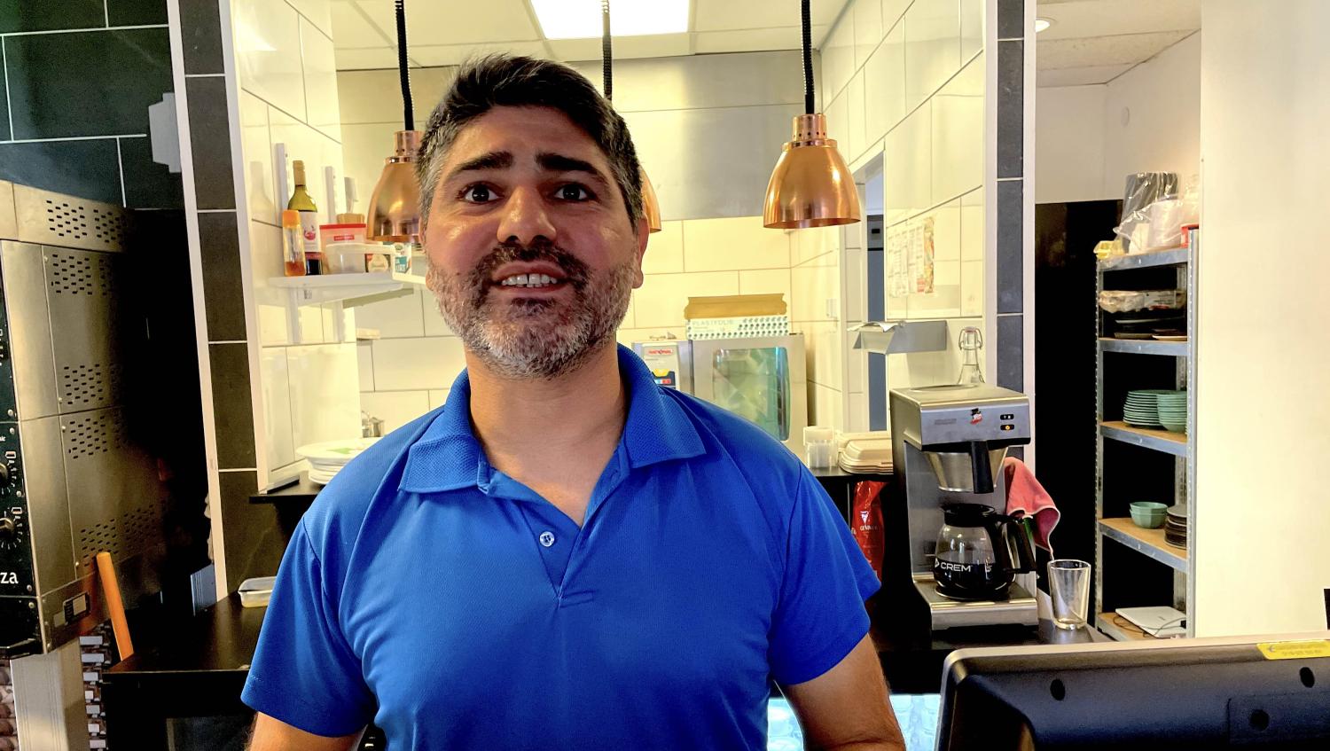 En man med kort hår och skägg, iklädd blå pikétröja, står bakom en disk på en restaurang. Köksredskap och buteljerade föremål syns i bakgrunden.