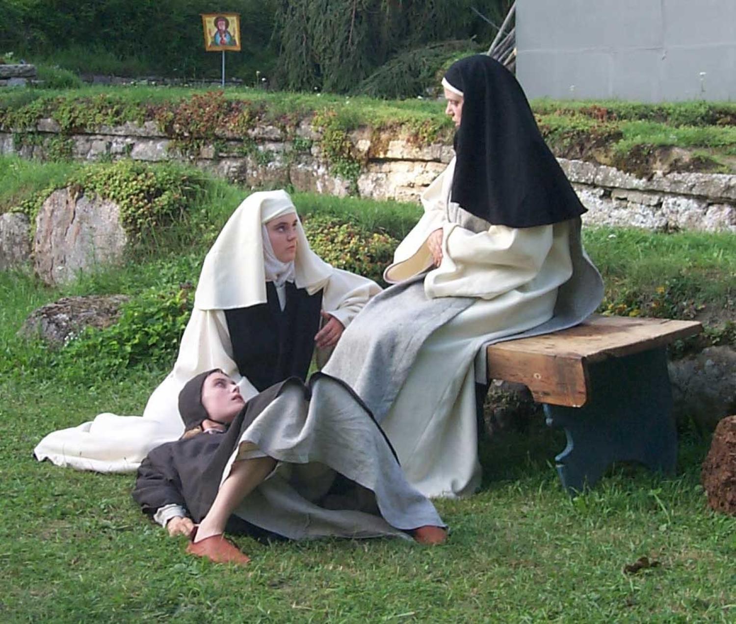 Tre nunnor i traditionella dräkter befinner sig utomhus; en sitter på en bänk, en sitter på marken och en annan ligger med huvudet i den andras knä. I bakgrunden syns en liten religiös ikon.