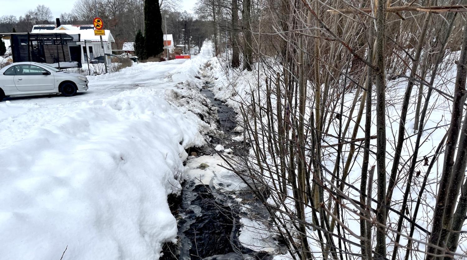 Snötäckt väg med parkerade bilar till vänster och en smal bäck med kala buskar till höger. Snöhögar kantar vägkanten.