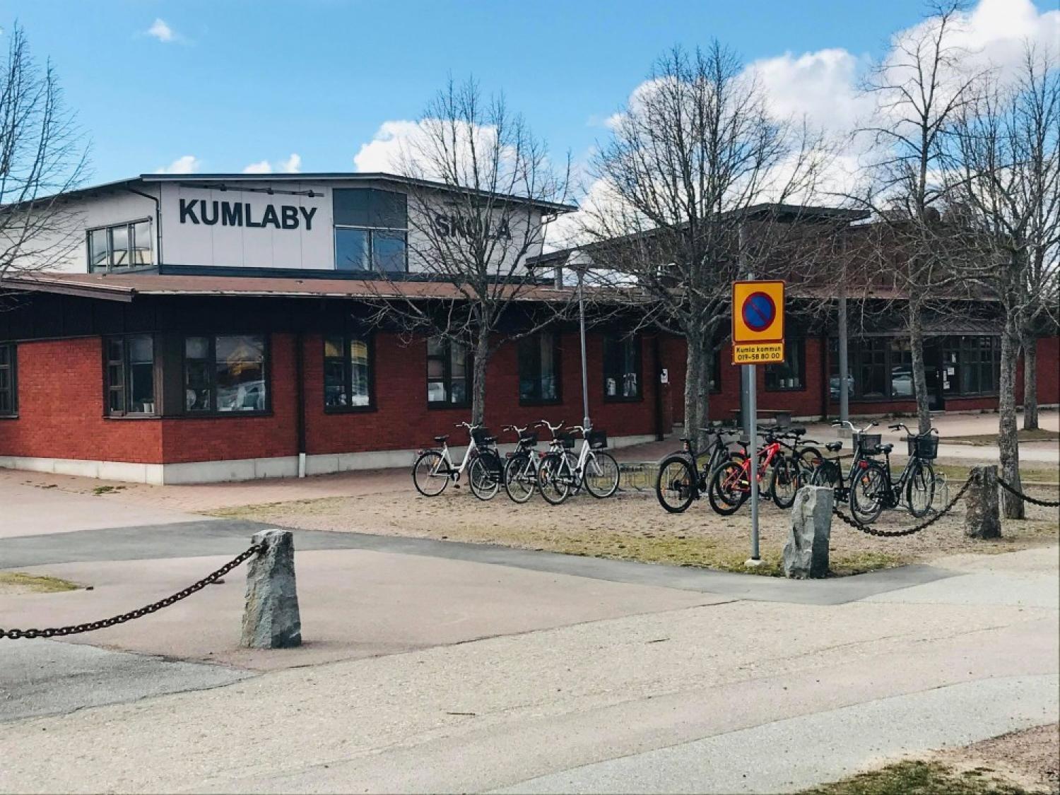 En skolbyggnad i rött tegel med skylten ”Kumlaby Skola”, med flera parkerade cyklar och lövlösa träd framför, under en delvis molnig himmel.