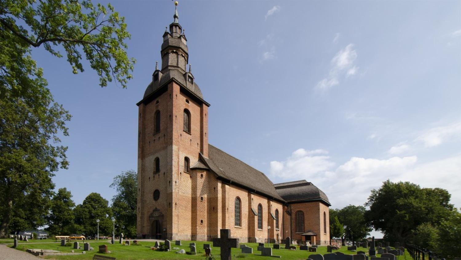 En stor tegelkyrka med ett högt fyrkantigt torn och välvda fönster, omgiven av en kyrkogård med gravstenar och grönt gräs under en delvis molnig himmel.