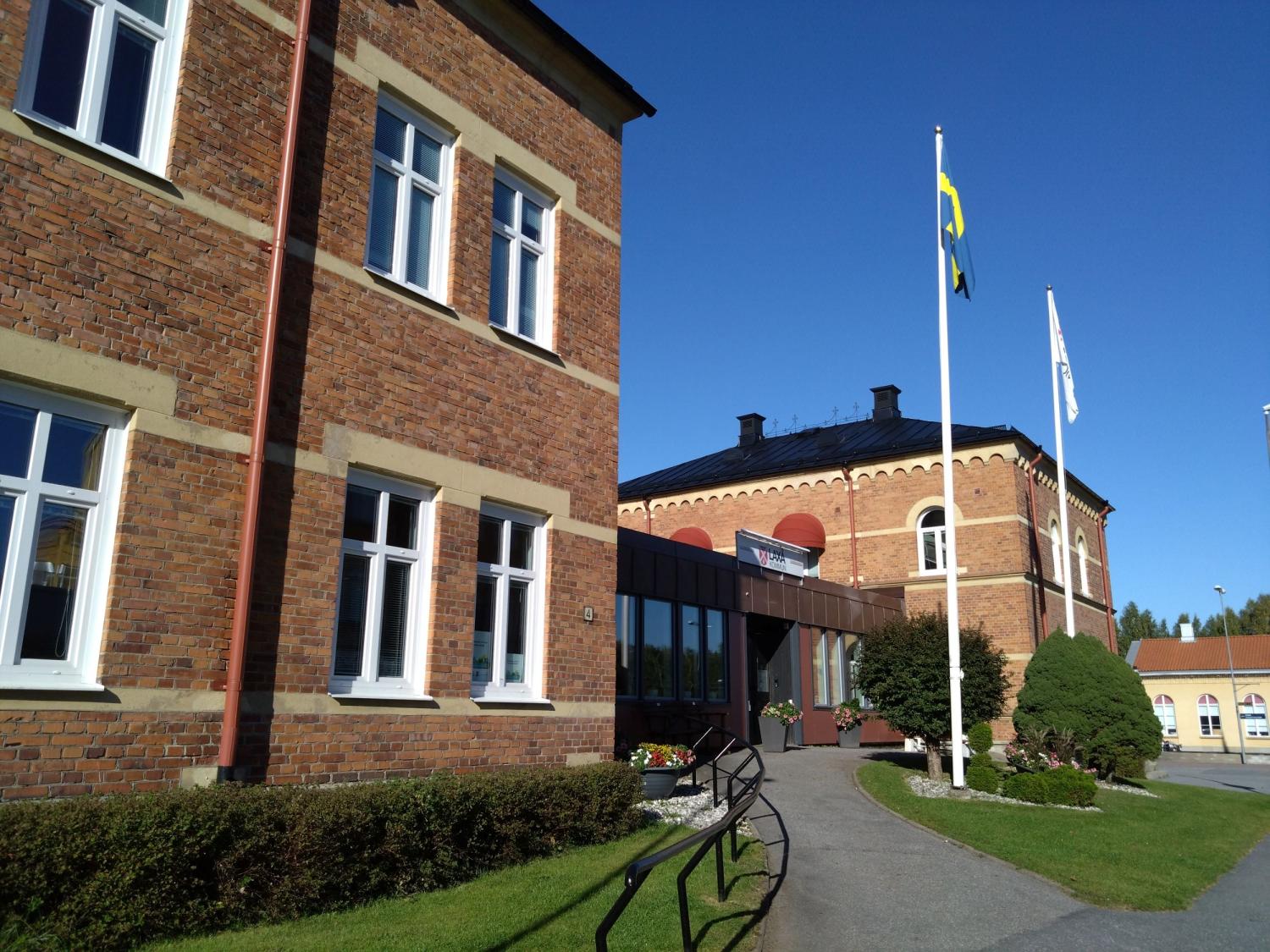 En tegelbyggnad med vita inramade fönster och en gångväg som leder till en entré. Två flaggstänger med flaggor står i närheten under en klarblå himmel.