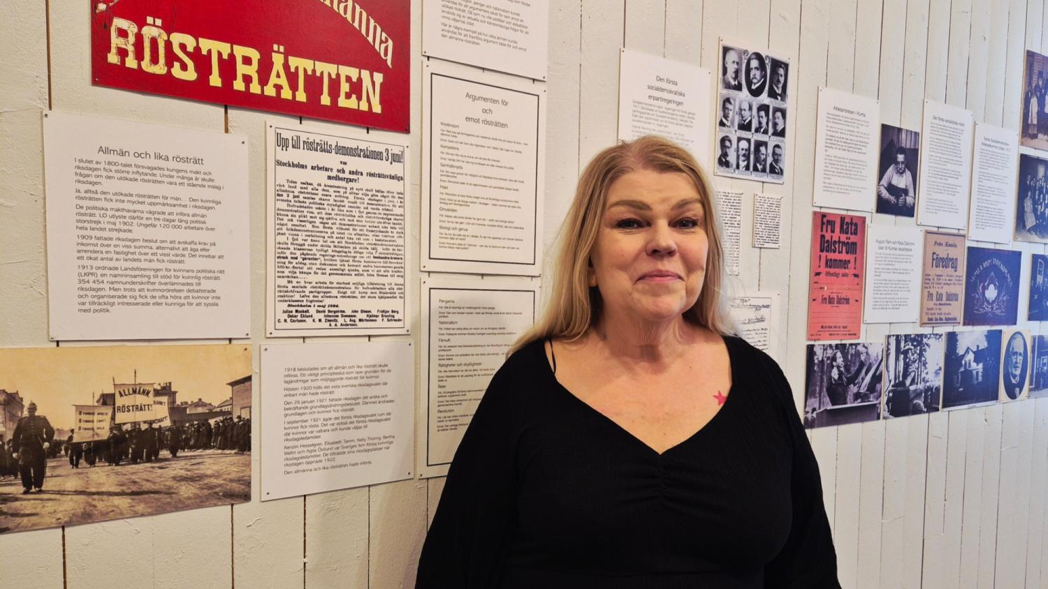En kvinna i svart topp står framför en vägg med historiska dokument, fotografier och affischer, bland annat en med svensk text om rösträtt.