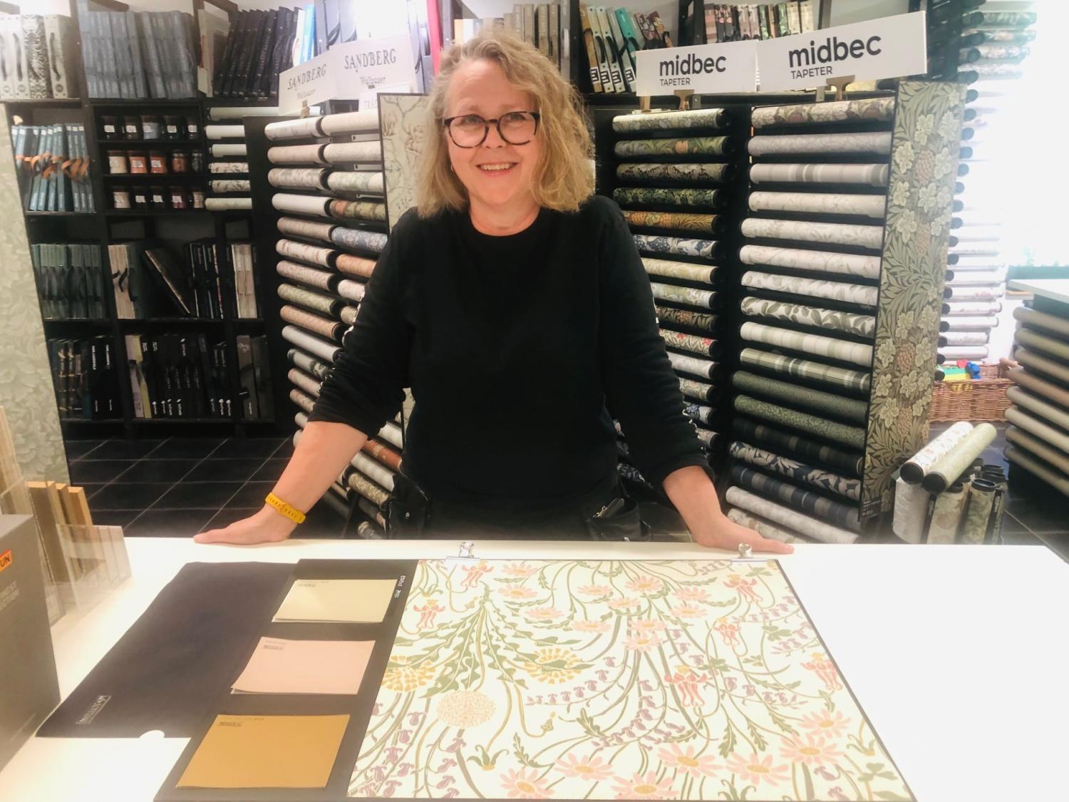 En person står bakom en disk och visar tapetprover och färgprover i en butik med hyllor med tapetrullar i bakgrunden.