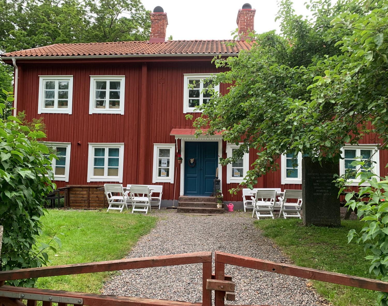 Ett traditionellt rött trähus i två våningar med vit dekor, en blå dörr, uteservering och grönska som omger trädgården och entrén.