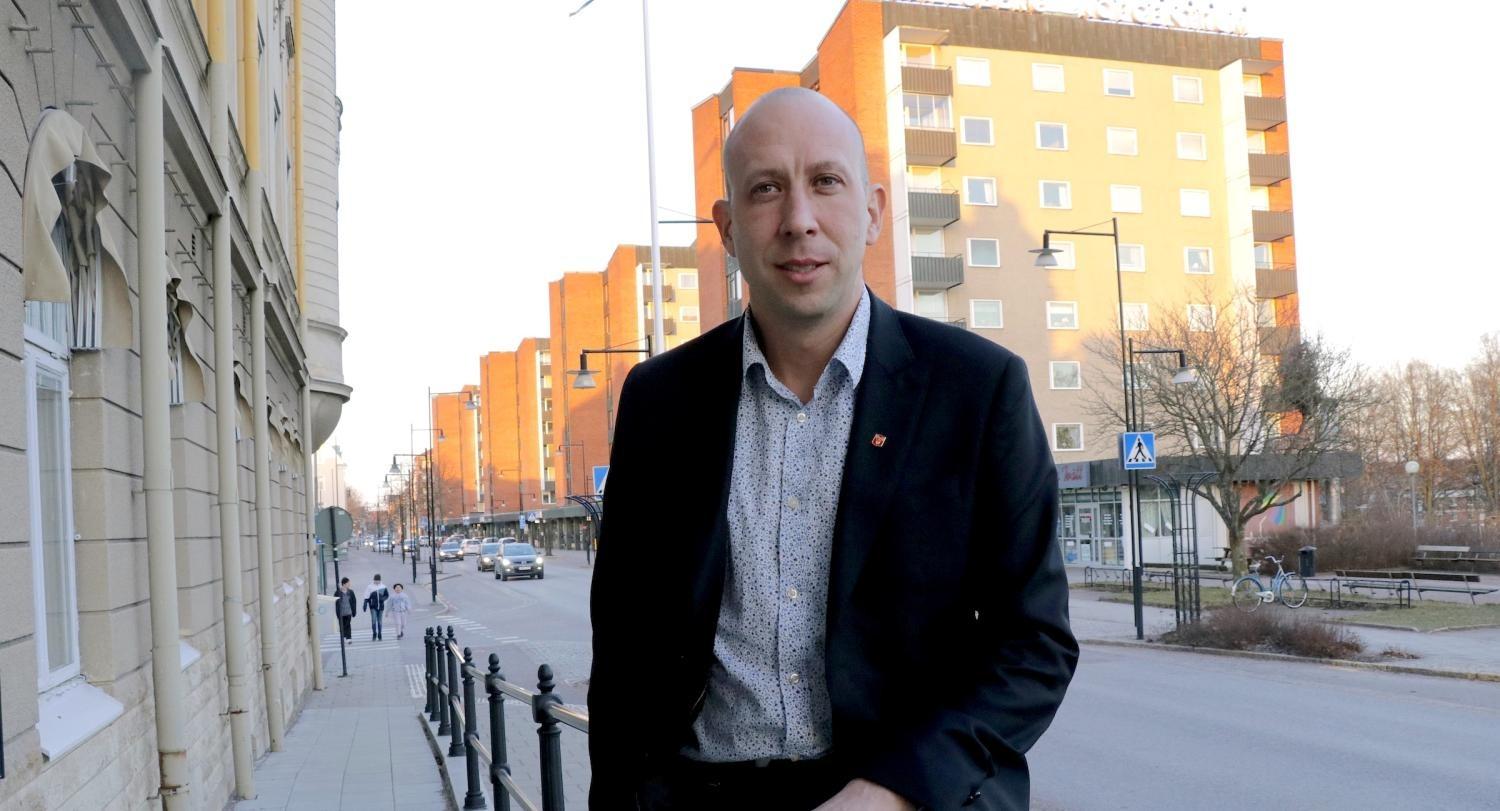 En man i kostym står på en trottoar i staden med flerbostadshus och några fotgängare i bakgrunden i dagsljus.