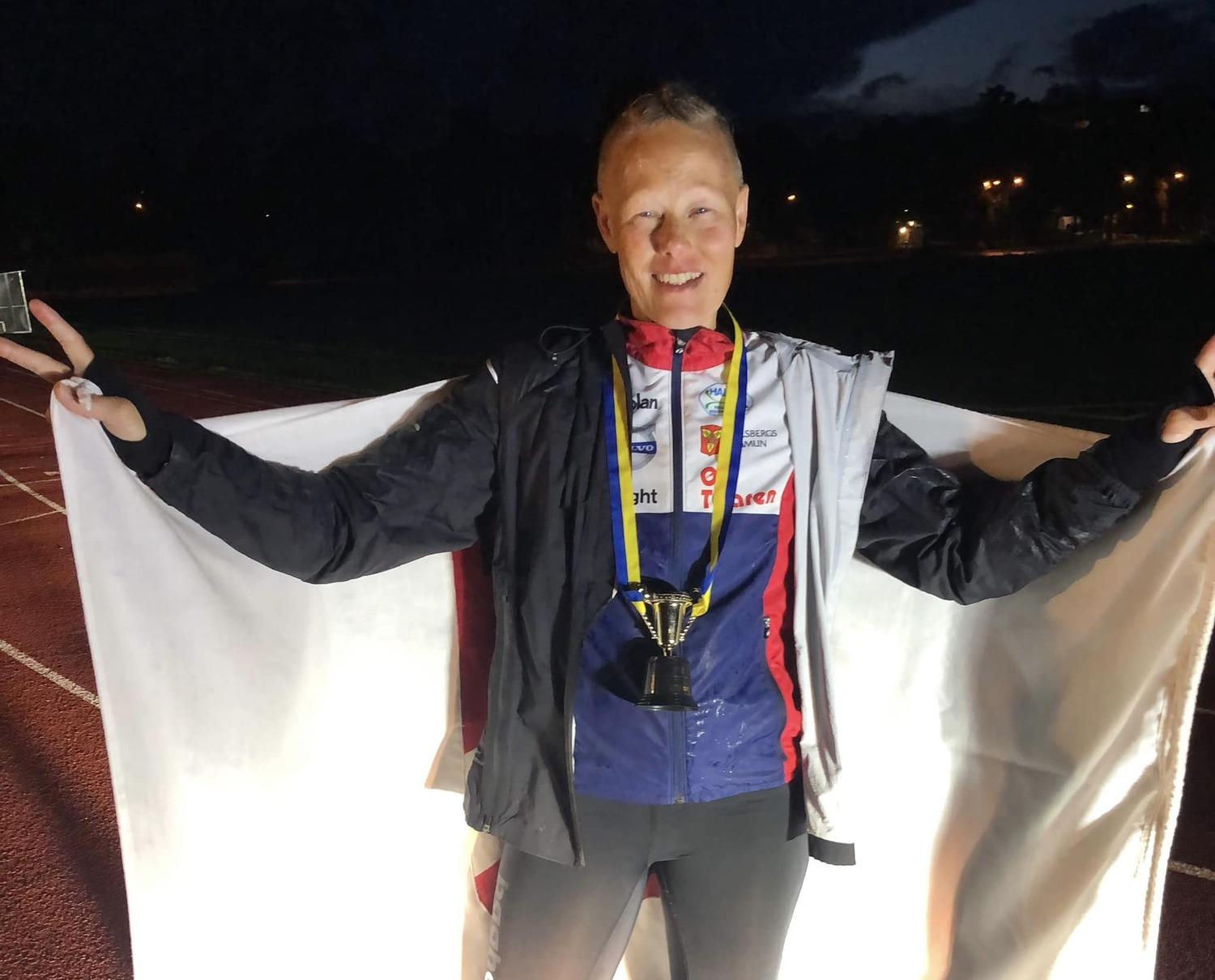 Person som står utomhus och håller en vit flagga, bär en medalj och ler. Det verkar vara mörkt eller sen kväll.