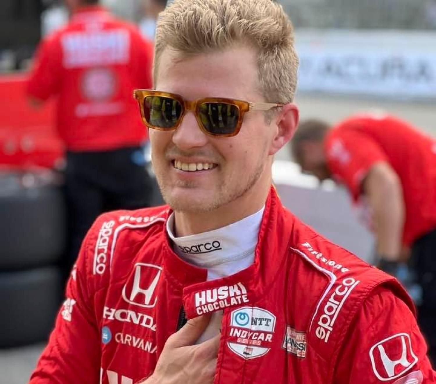 En racerbilsförare i röd racingdräkt och solglasögon ler när han står utomhus tillsammans med andra teammedlemmar i matchande röda kläder.