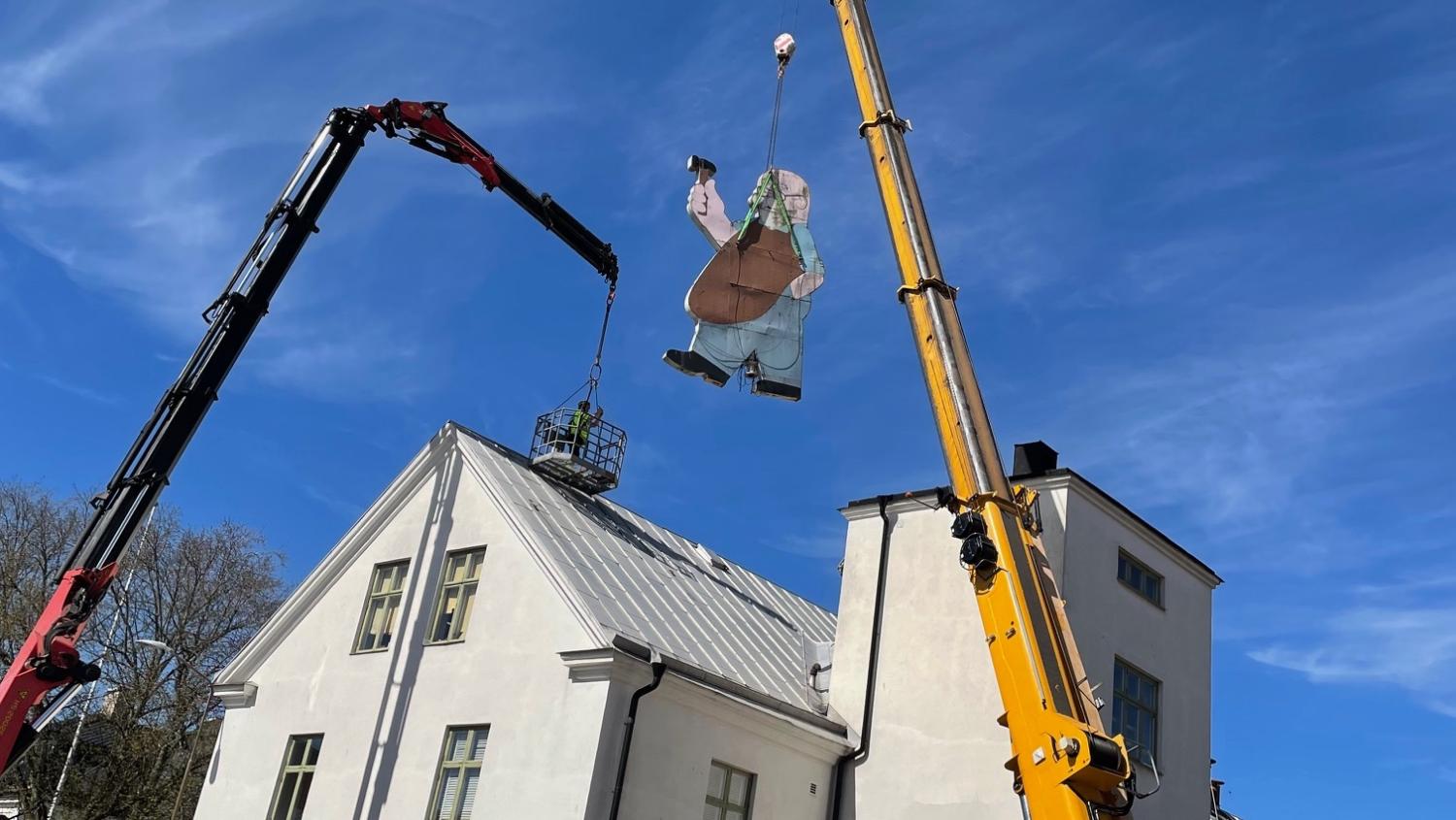 Två lyftkranar lyfter en stor platt utskärning av en person över taket på en vit byggnad en klar, solig dag.