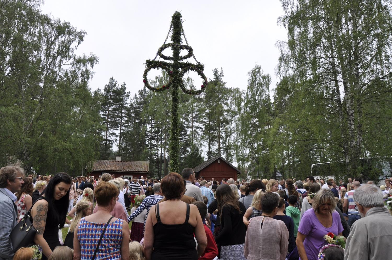 Skara människor som firar en traditionell svensk midsommarfest utomhus, med en stor majstång dekorerad med löv och blommor i bakgrunden.