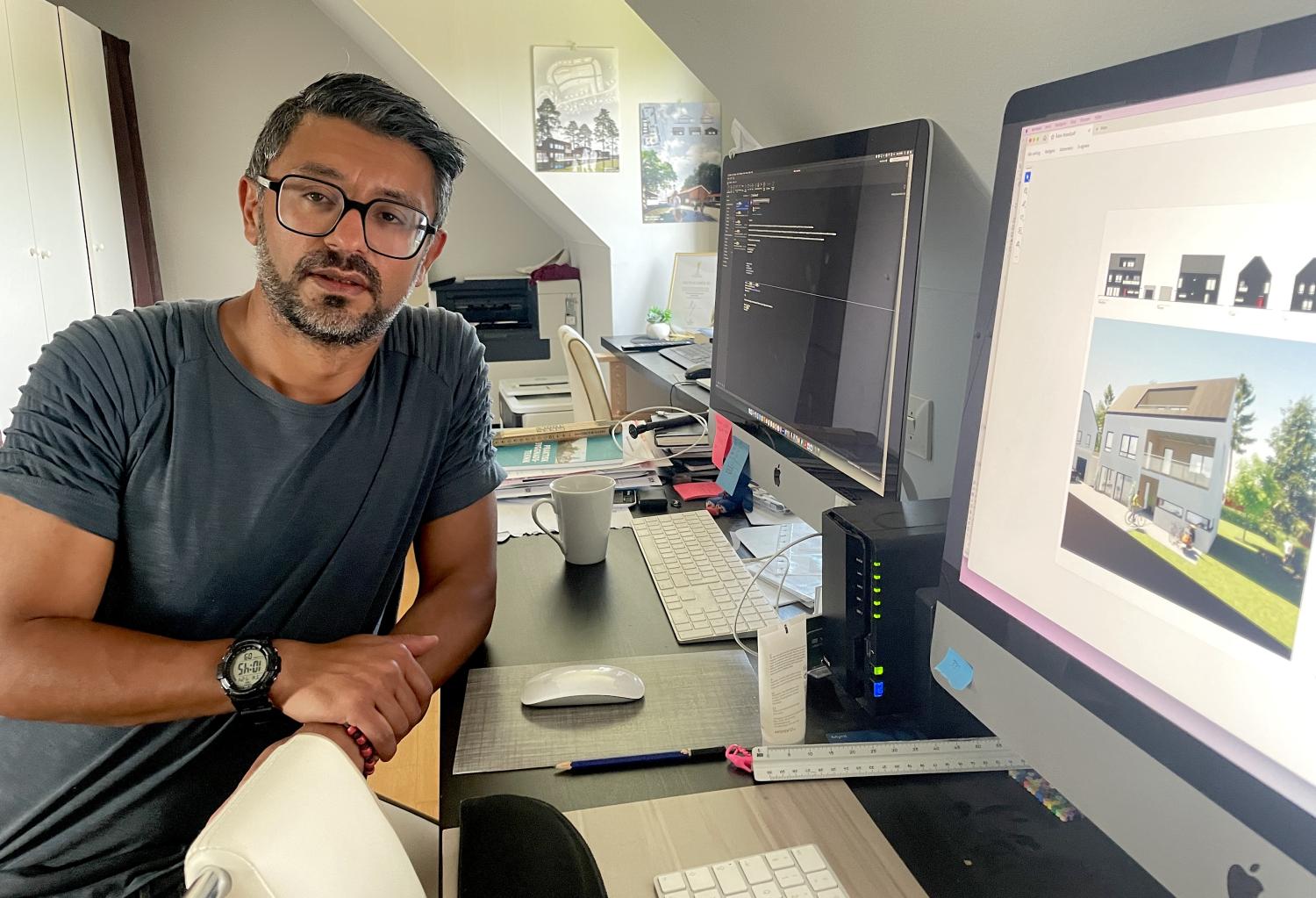 En man står vid ett skrivbord med två skärmar, den ena visar kod och den andra visar arkitektoniska renderingar. På skrivbordet finns diverse kontorsmaterial och en kaffemugg.