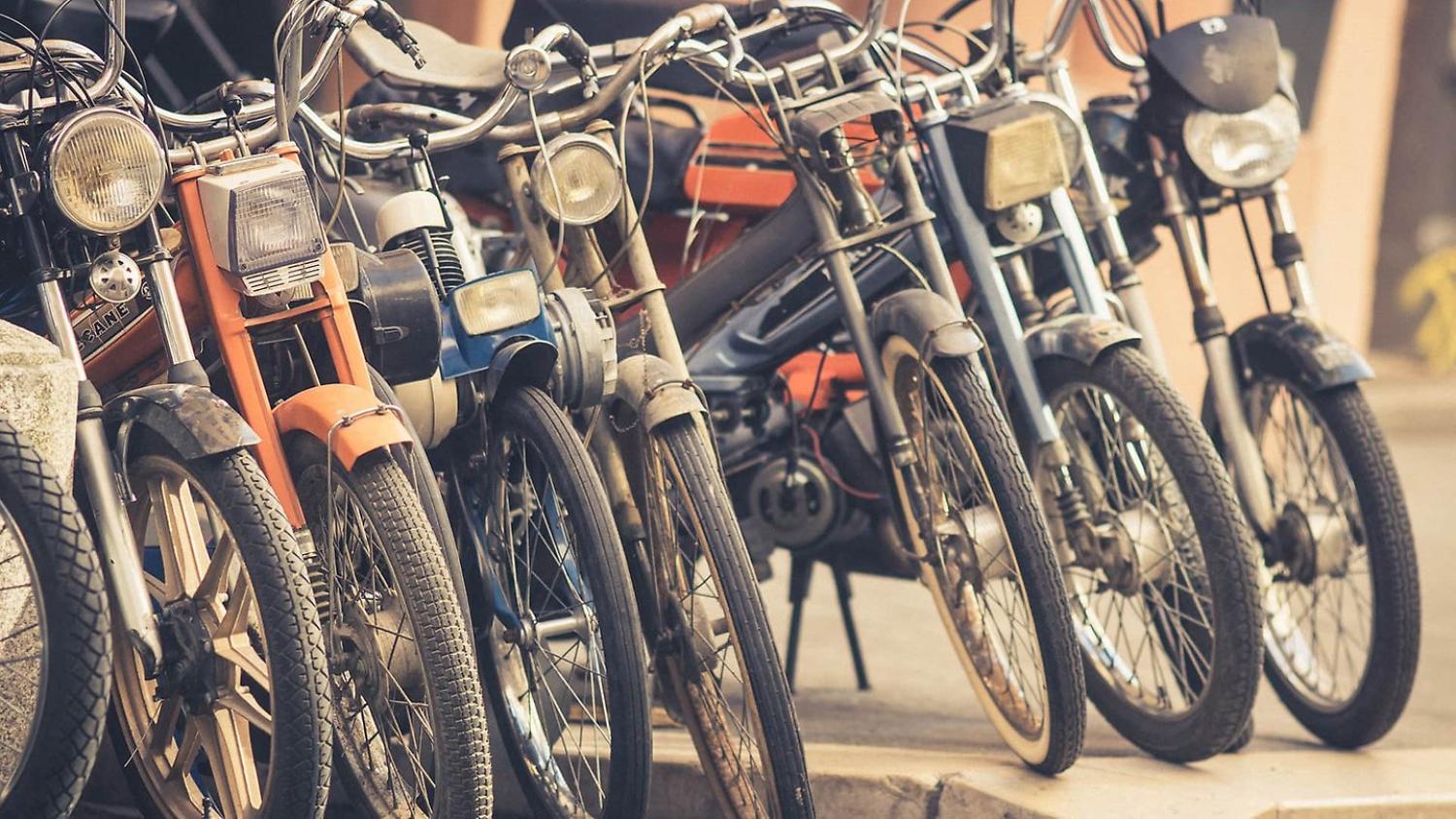 En rad veteranmopeder och cyklar står tätt parkerade på en trottoar.