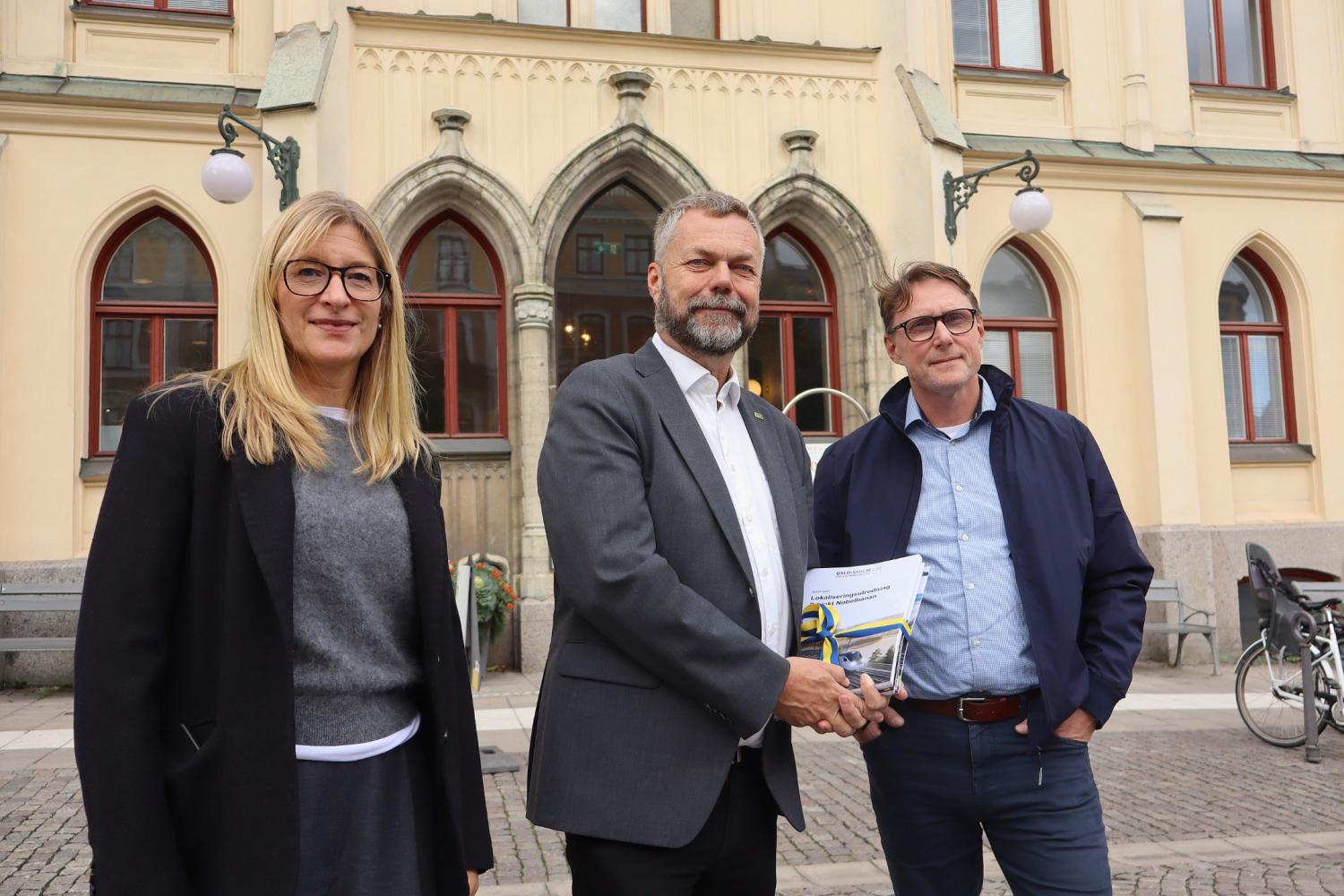 Tre vuxna står utanför en historisk byggnad; en man i kostym håller i några dokument, med en kvinna och en annan man på vardera sidan om sig.