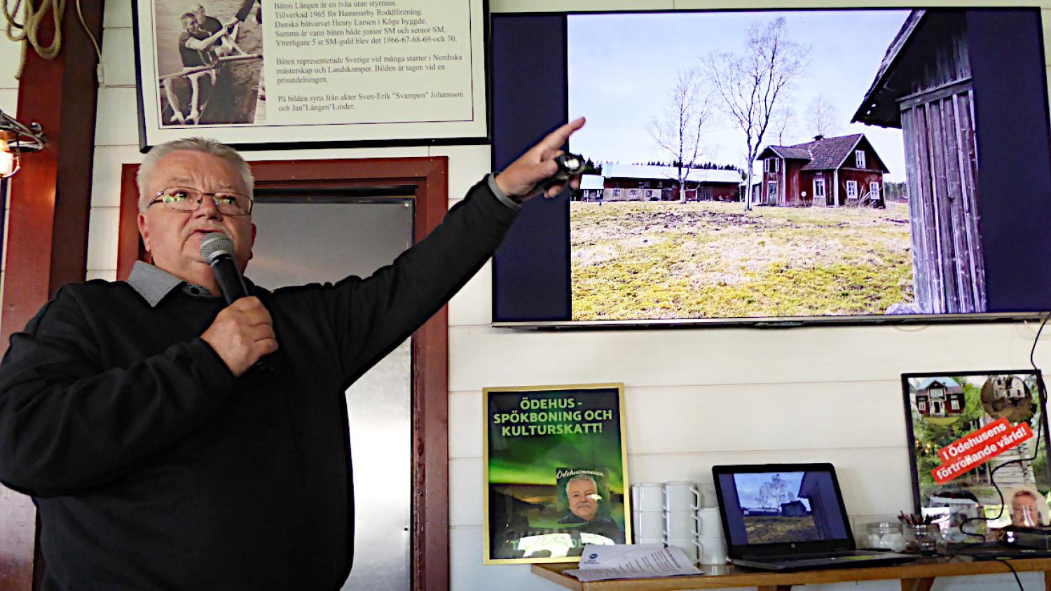 En äldre man med mikrofon pekar på en skärm som visar en husmiljö på landsbygden; bredvid honom syns informationsaffischer och ett litet bord.