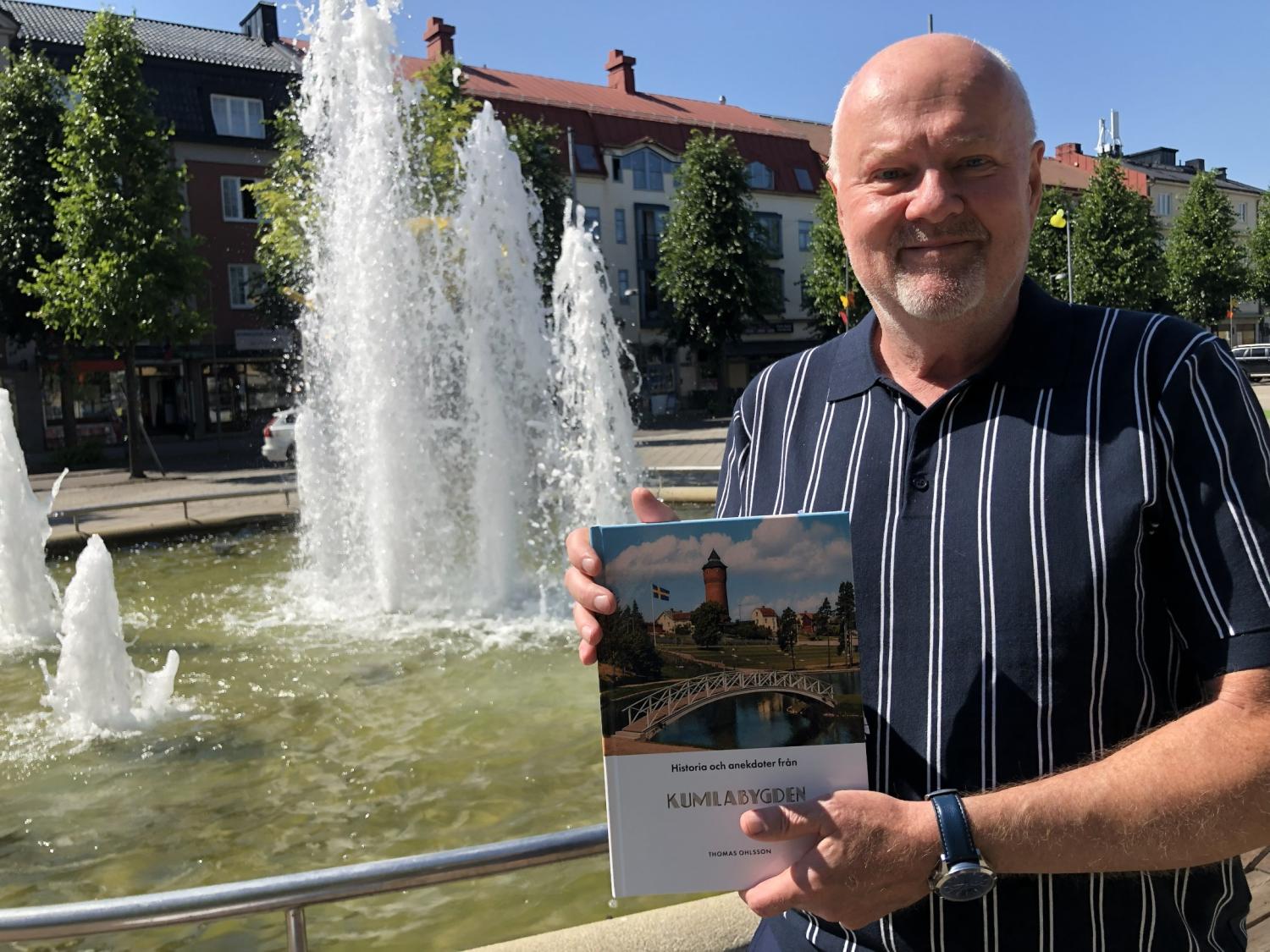 En man står framför en fontän och håller i en bok med titeln ”Kumlabygden”. I bakgrunden syns byggnader och träd.
