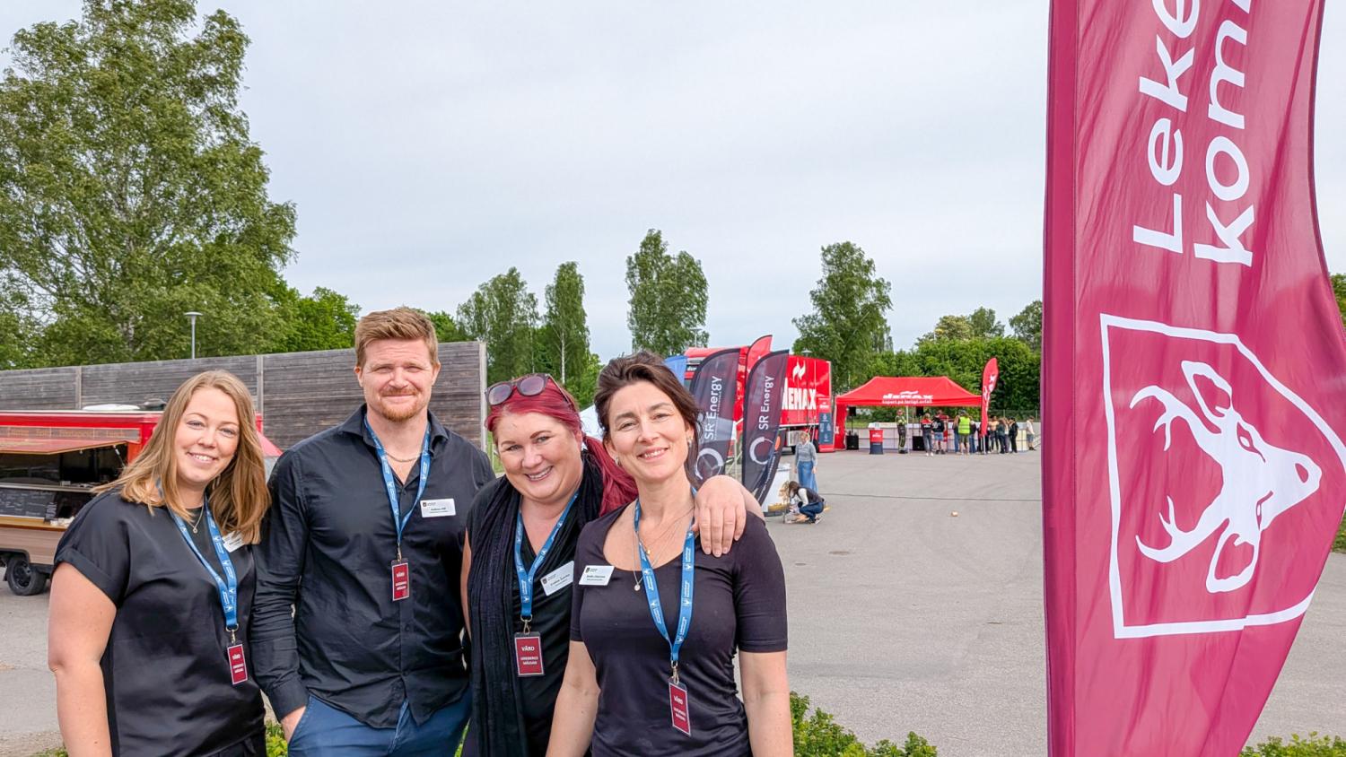 Fyra personer med namnbrickor och nyckelband står utomhus nära en stor flagga med en tjurlogotyp och text. Röda tält och träd syns i bakgrunden.