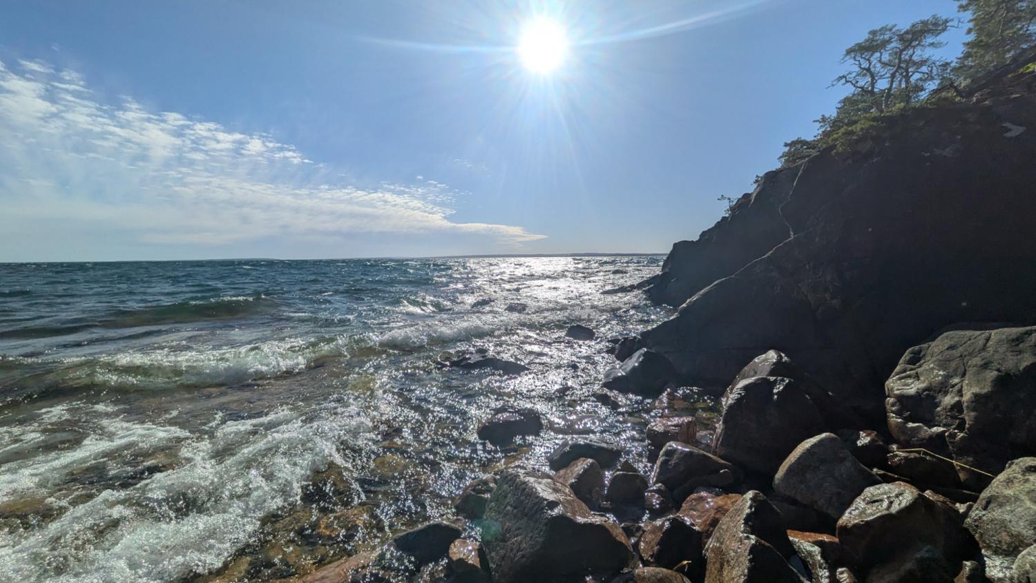 Klippig strandlinje med vågor som slår in under en stark sol, en delvis molnig himmel och träd på en klippa på höger sida.