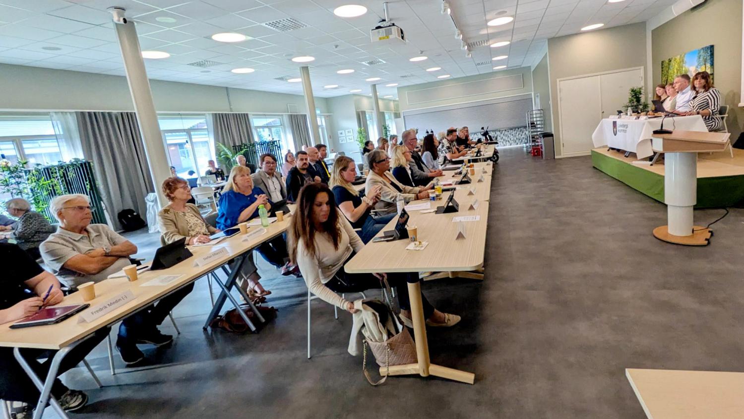Personer som sitter vid bord deltar i ett möte eller en konferens i en modern lokal, medan fyra paneldeltagare talar på scenen vid ett bord.