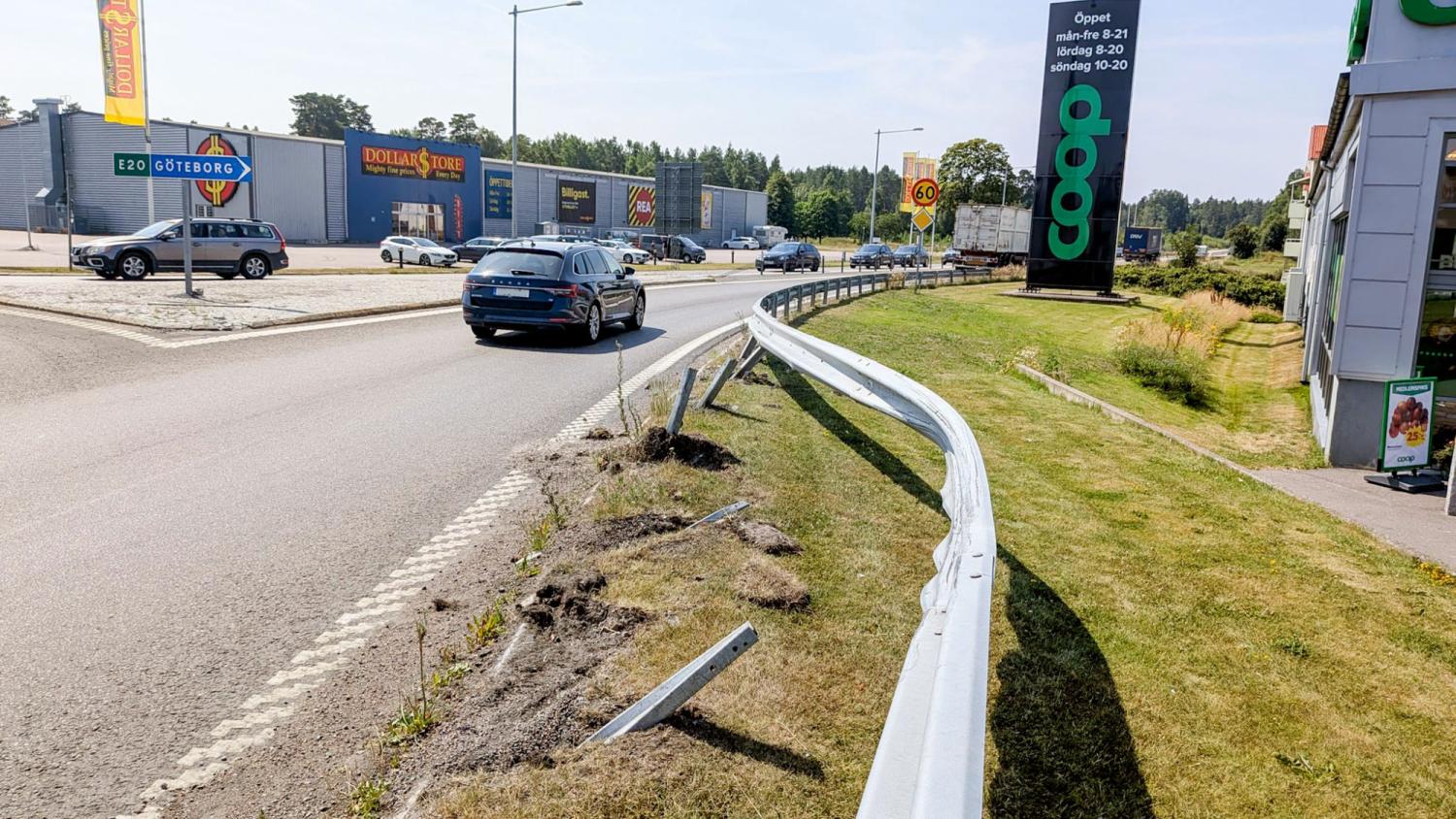 Ett skadat vägräcke med böjda delar löper längs en trafikerad gata nära butiker, en parkeringsplats och en skylt som visar Coop-supermarknadens öppettider.