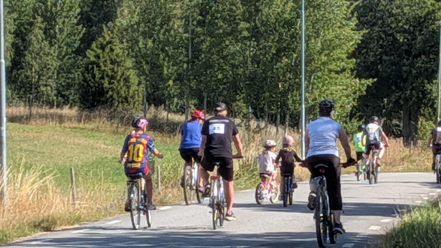 En grupp människor, bestående av både vuxna och barn, cyklar tillsammans på en asfalterad väg som kantas av gräs och träd.