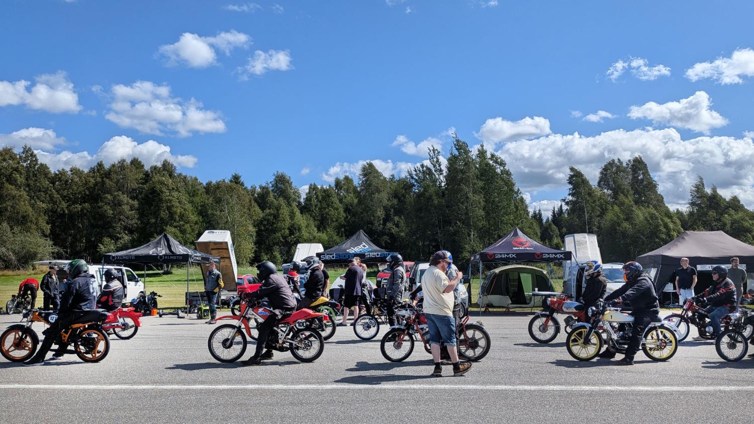 Människor på små motorcyklar står uppställda på en väg nära flera tält, med åskådare och träd i bakgrunden under en delvis molnig himmel.