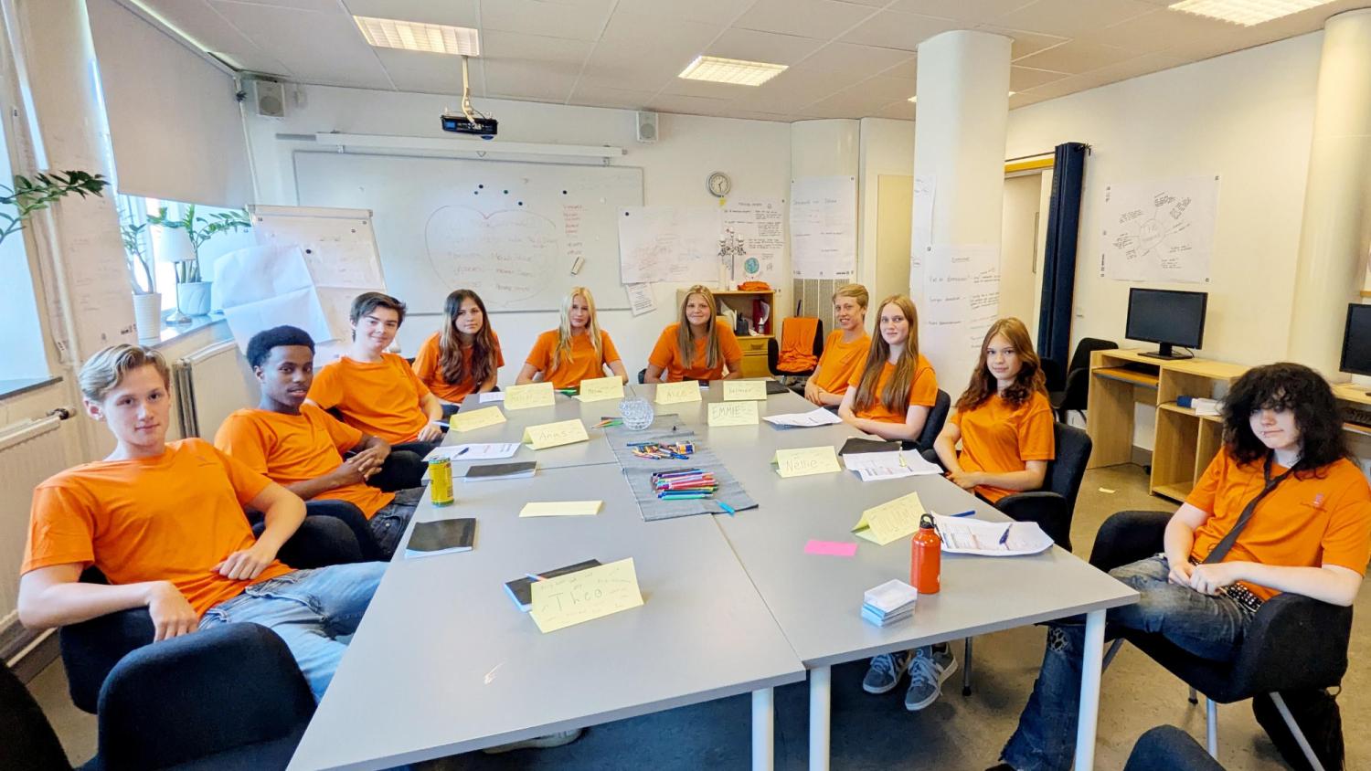 Tolv ungdomar i orangea tröjor sitter runt ett bord i ett klassrum med papper, namnskyltar och tuschpennor, vända mot kameran.